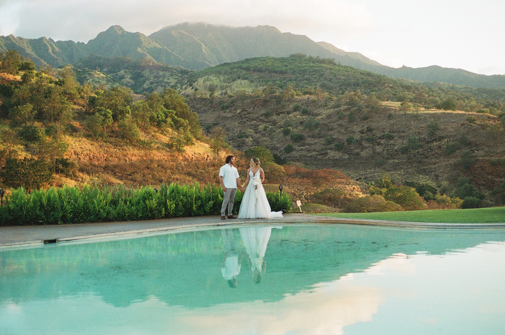 Bride and groom standing hand in hand near a pool reflecting the mountains and golden sky at a dreamy Wedding Venues in Hawaii.