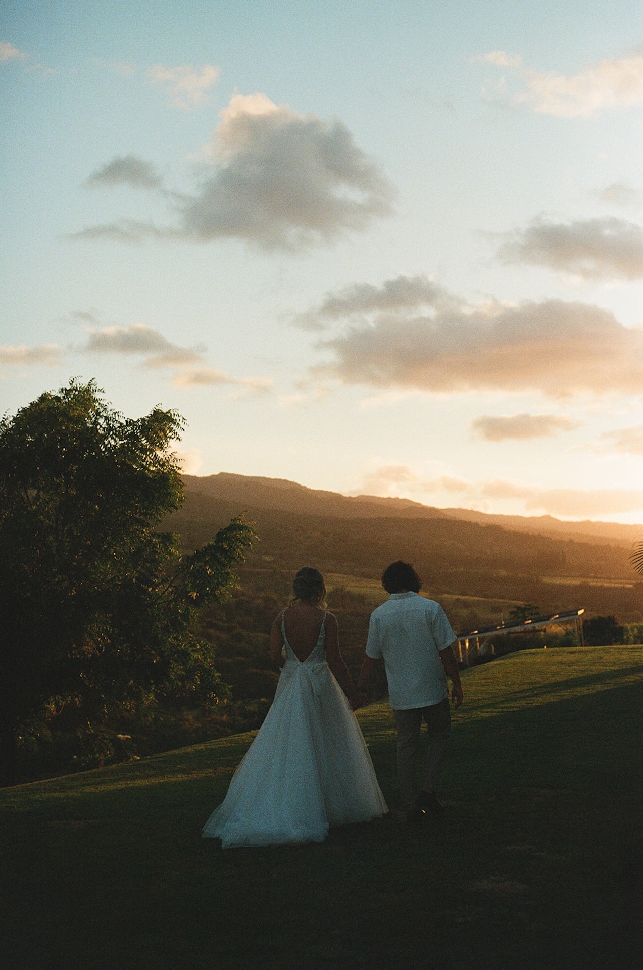Bride and groom walking hand in hand down a grassy hill at sunset, their silhouettes glowing against the sky.