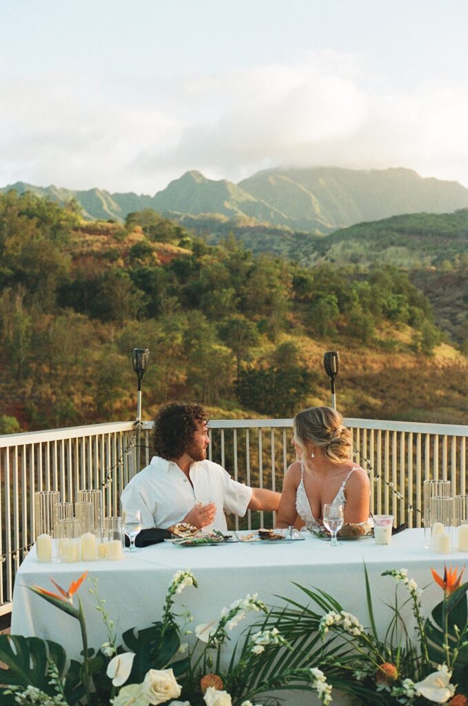 Bride and groom sharing dinner together on a mountaintop patio with tropical flowers and scenic views at sunset.