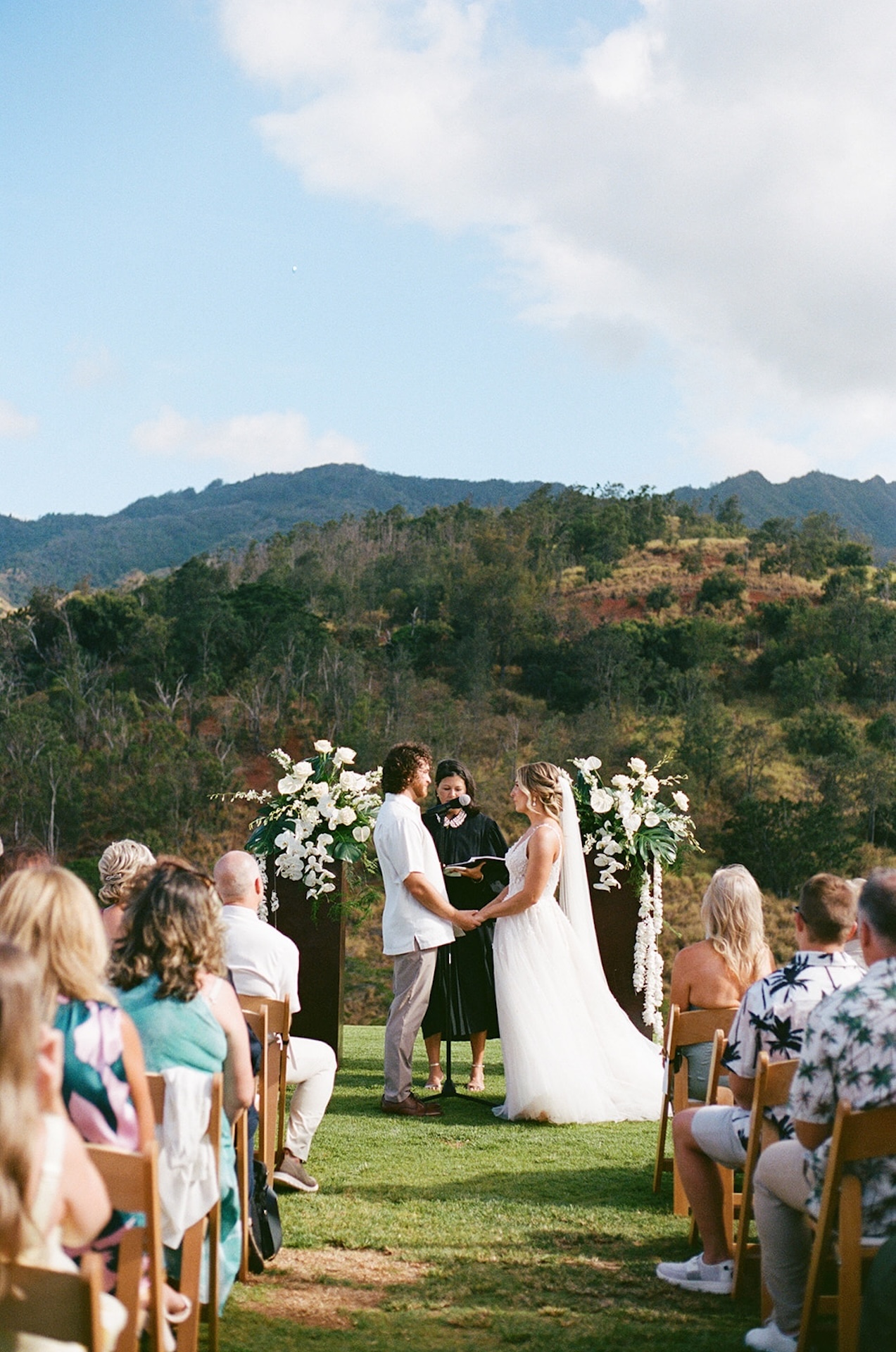 Bride and groom holding hands at the altar with the officiant between them, surrounded by guests and panoramic mountain views at a stunning Wedding Venues in Hawaii.