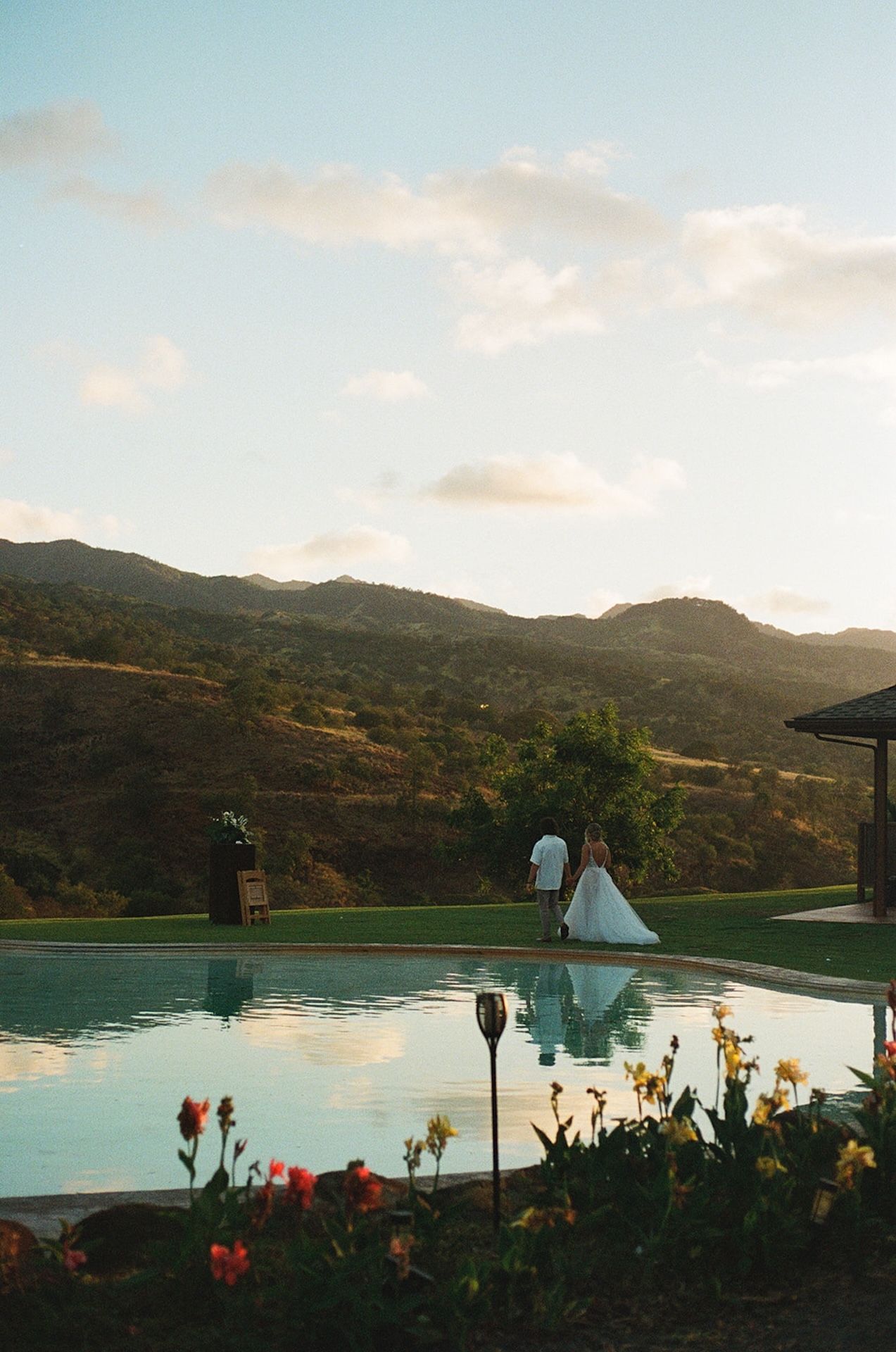Couple walking near a pool reflecting the golden hour sky, with colorful flowers in the foreground and peaceful mountain views.