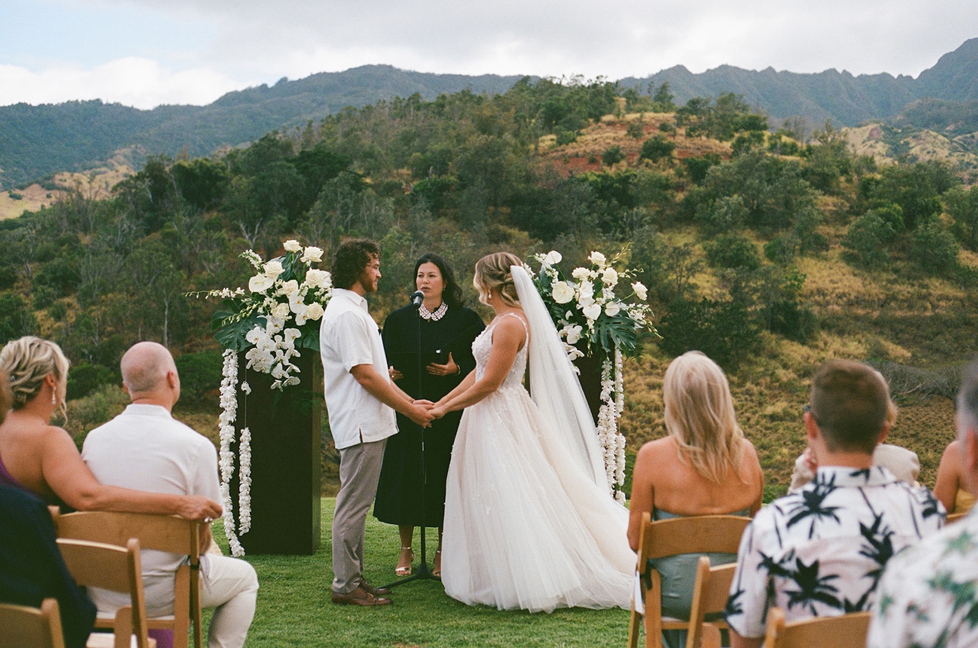 Bride and groom holding hands during the ceremony with floral pillars, mountains in the distance, and guests watching at a scenic Wedding Venues in Hawaii.