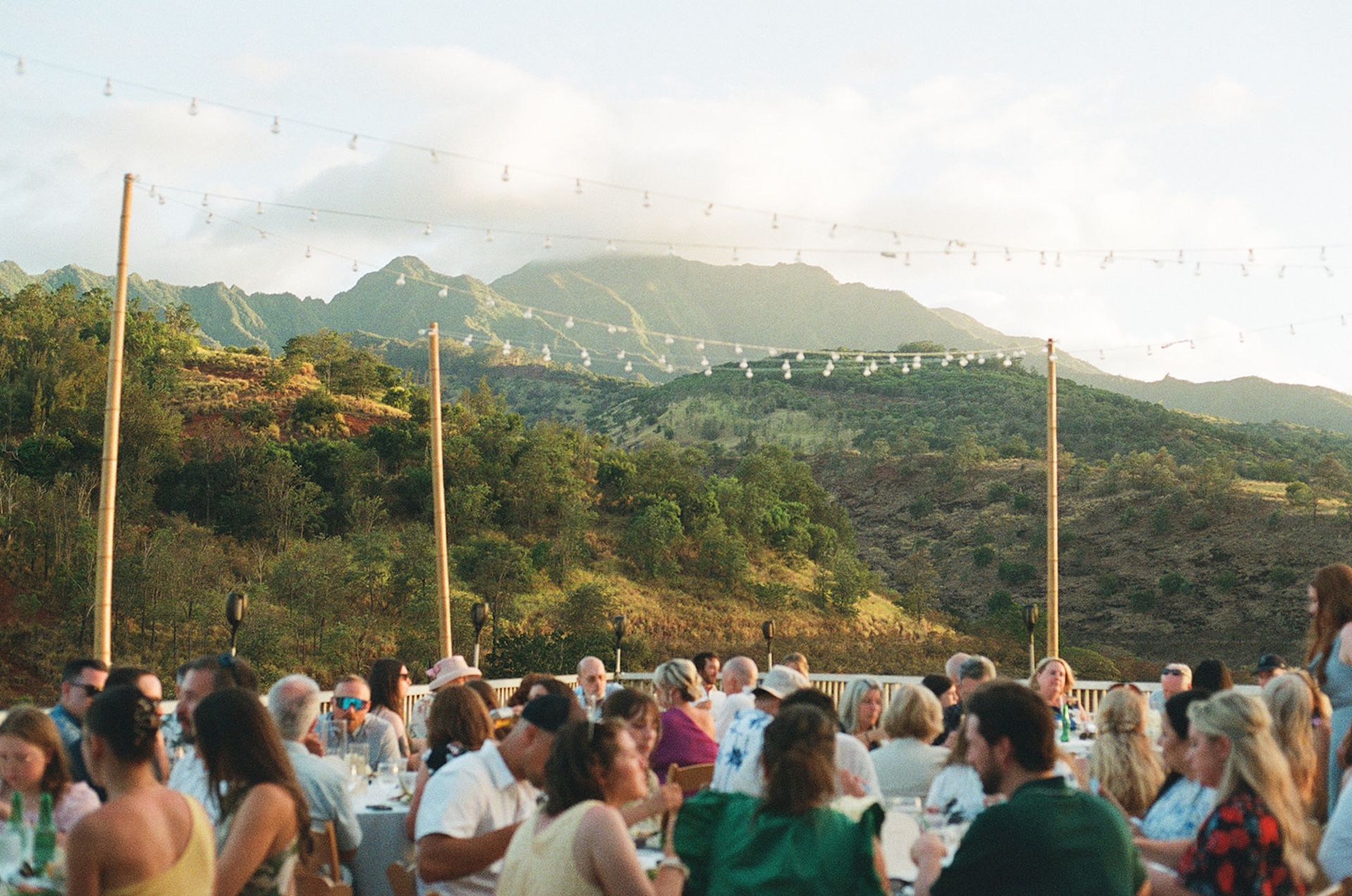 Guests seated at long tables enjoying dinner outdoors with the mountains glowing in golden light, a dreamy view of Wedding Venues in Hawaii.