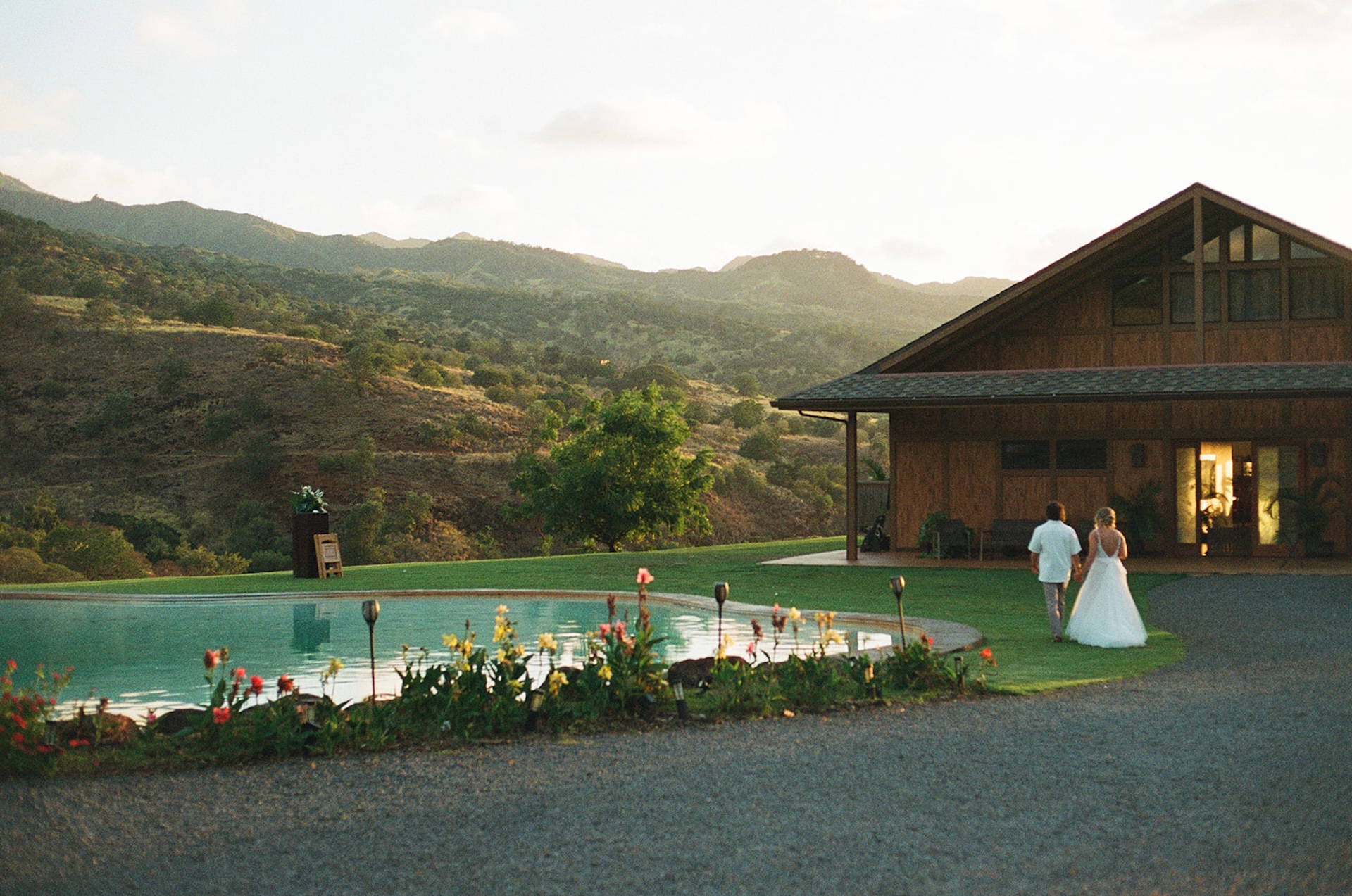 Couple walking back toward a wooden lodge near the pool as the sun sets behind the hills, a quiet and intimate end to their day.