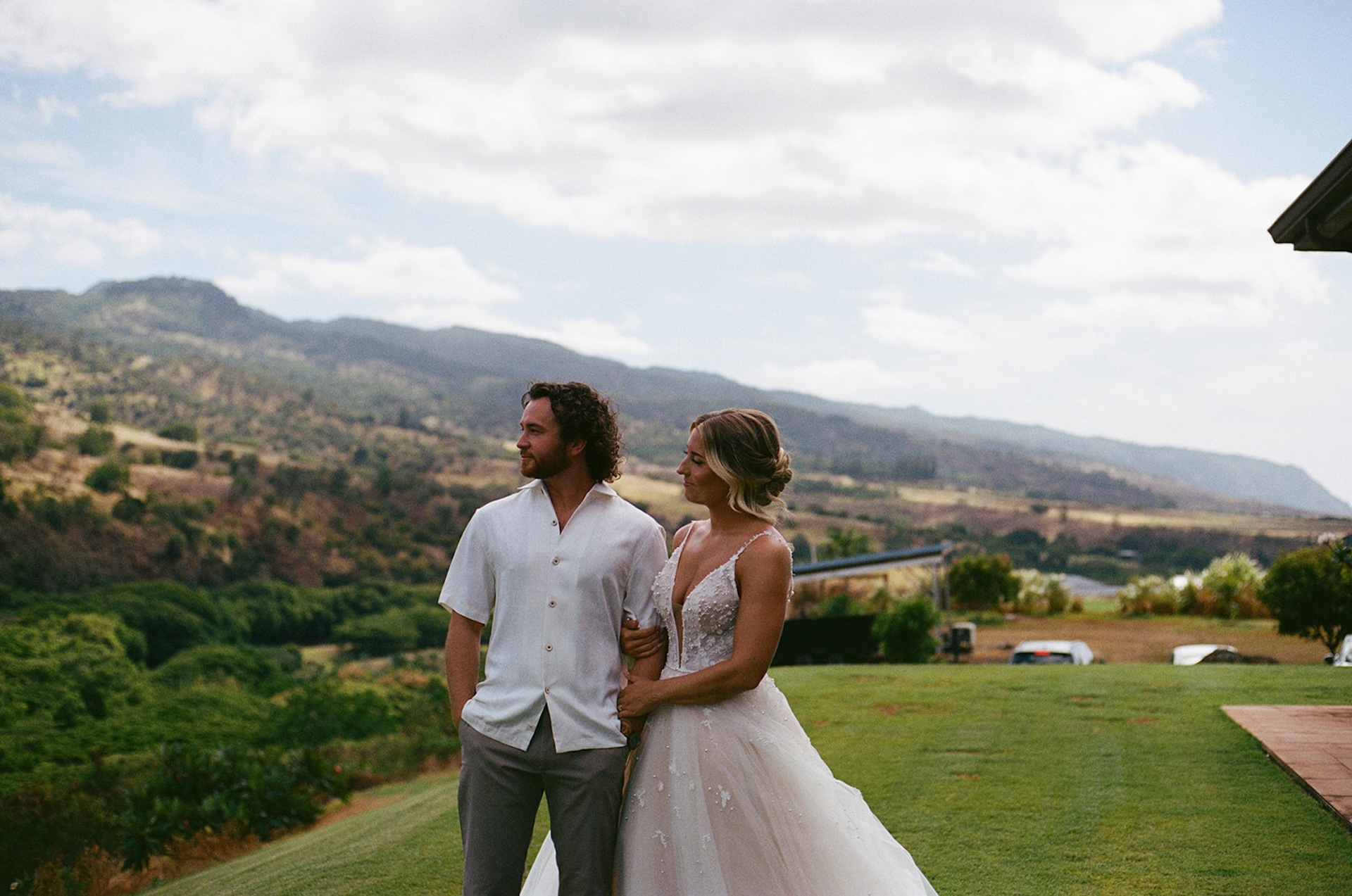 Bride and groom standing arm in arm overlooking lush green mountains and valleys under a partly cloudy sky.