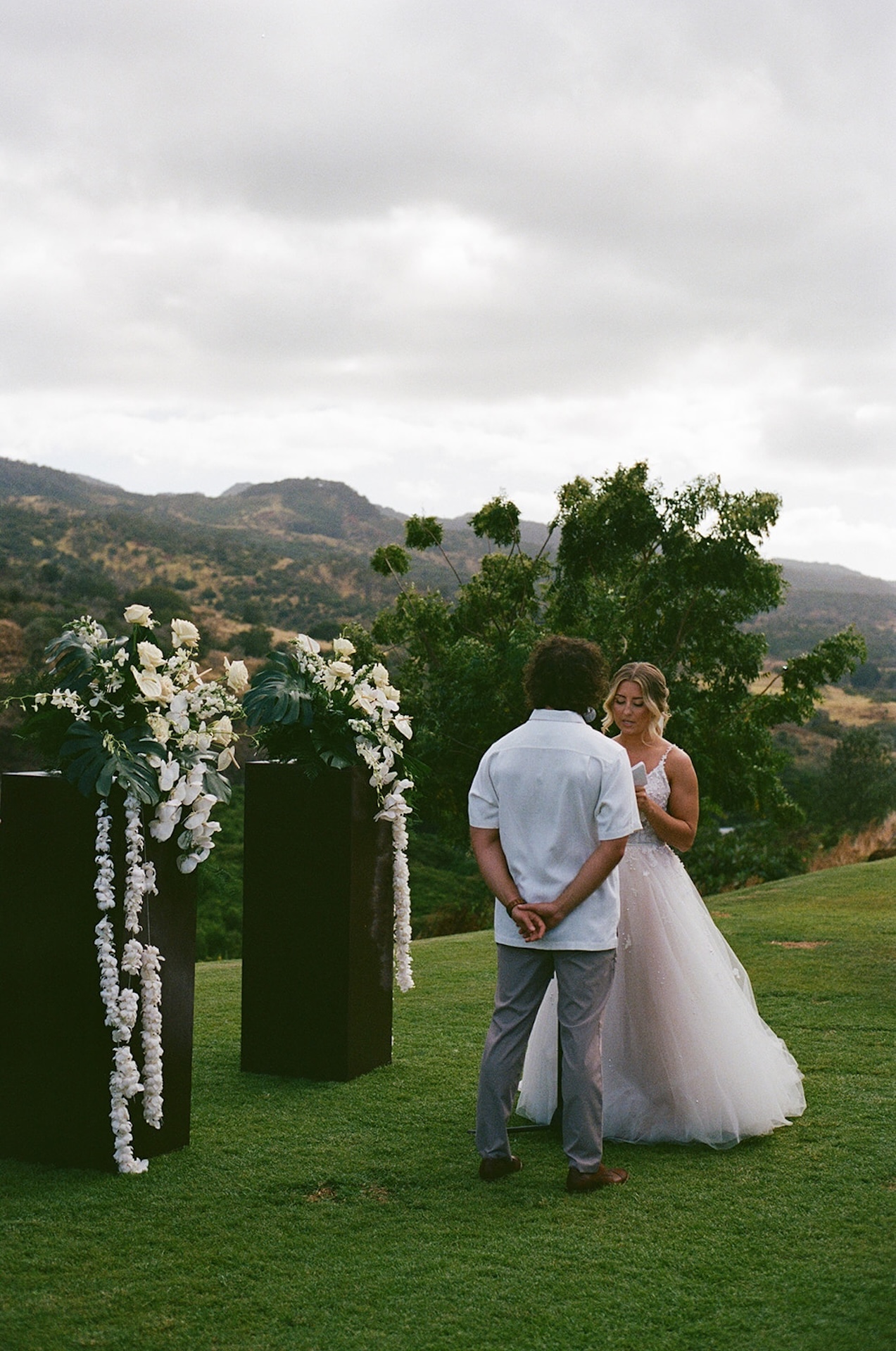 Bride reading vows to her groom on a grassy hilltop with elegant floral pillars and overcast skies.