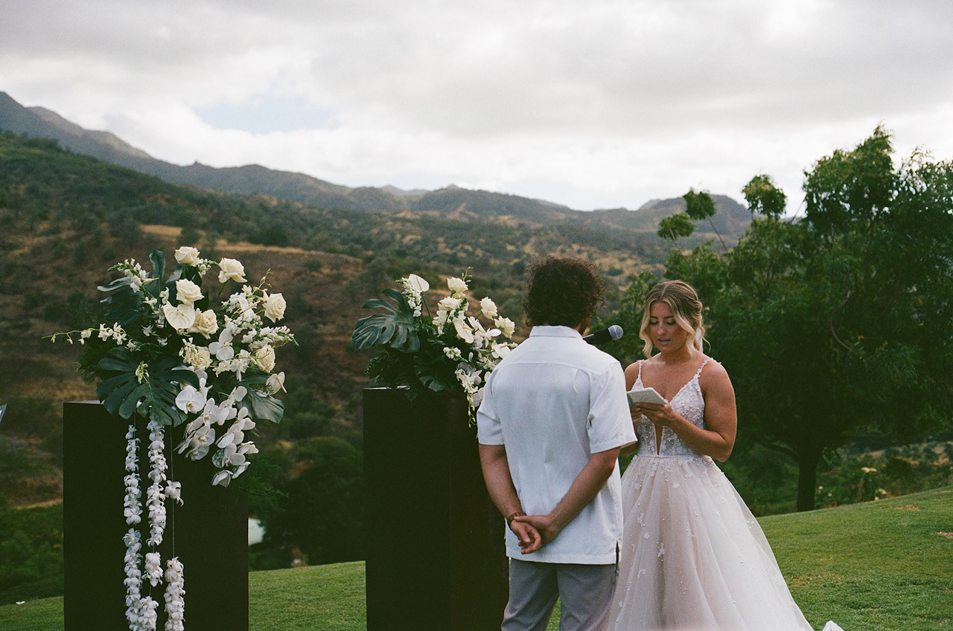 Bride reading her vows to the groom outdoors surrounded by lush greenery and tropical floral arrangements.