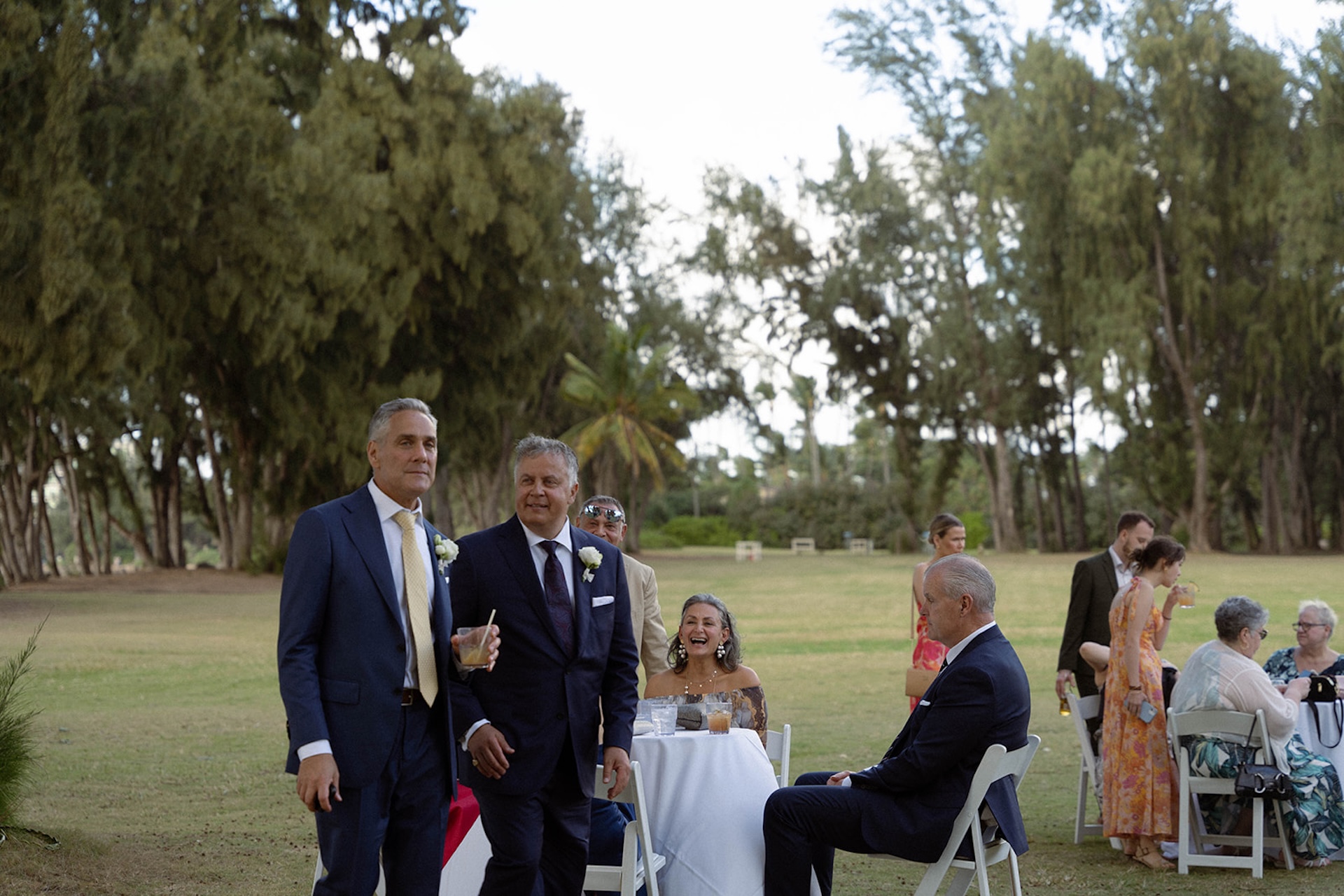 Two groomsmen in suits walk into cocktail hour smiling, as guests laugh and enjoy drinks at outdoor tables.
