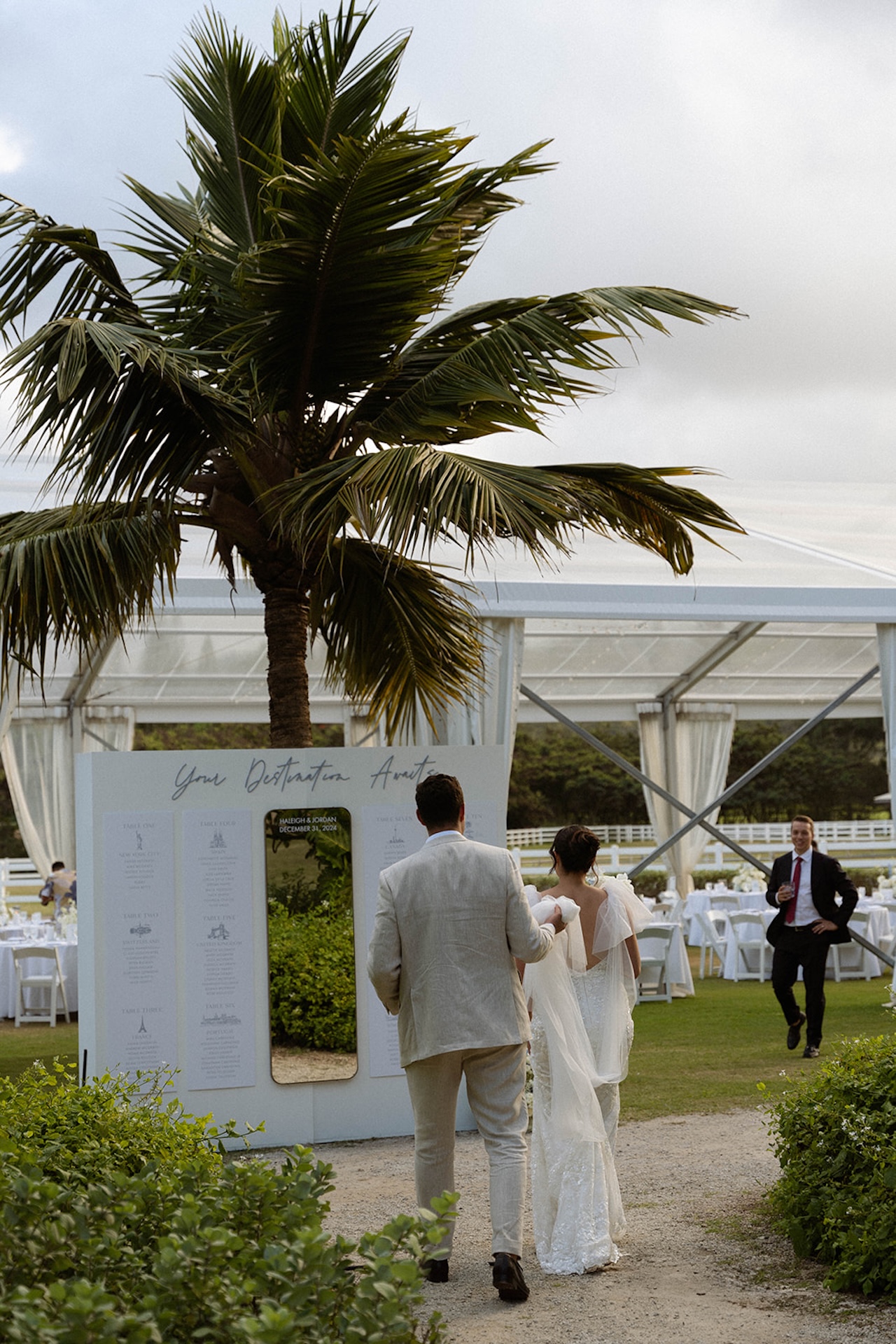 The couple walks hand in hand toward their seating chart display framed by a palm tree, entering the reception tent as guests cheer.