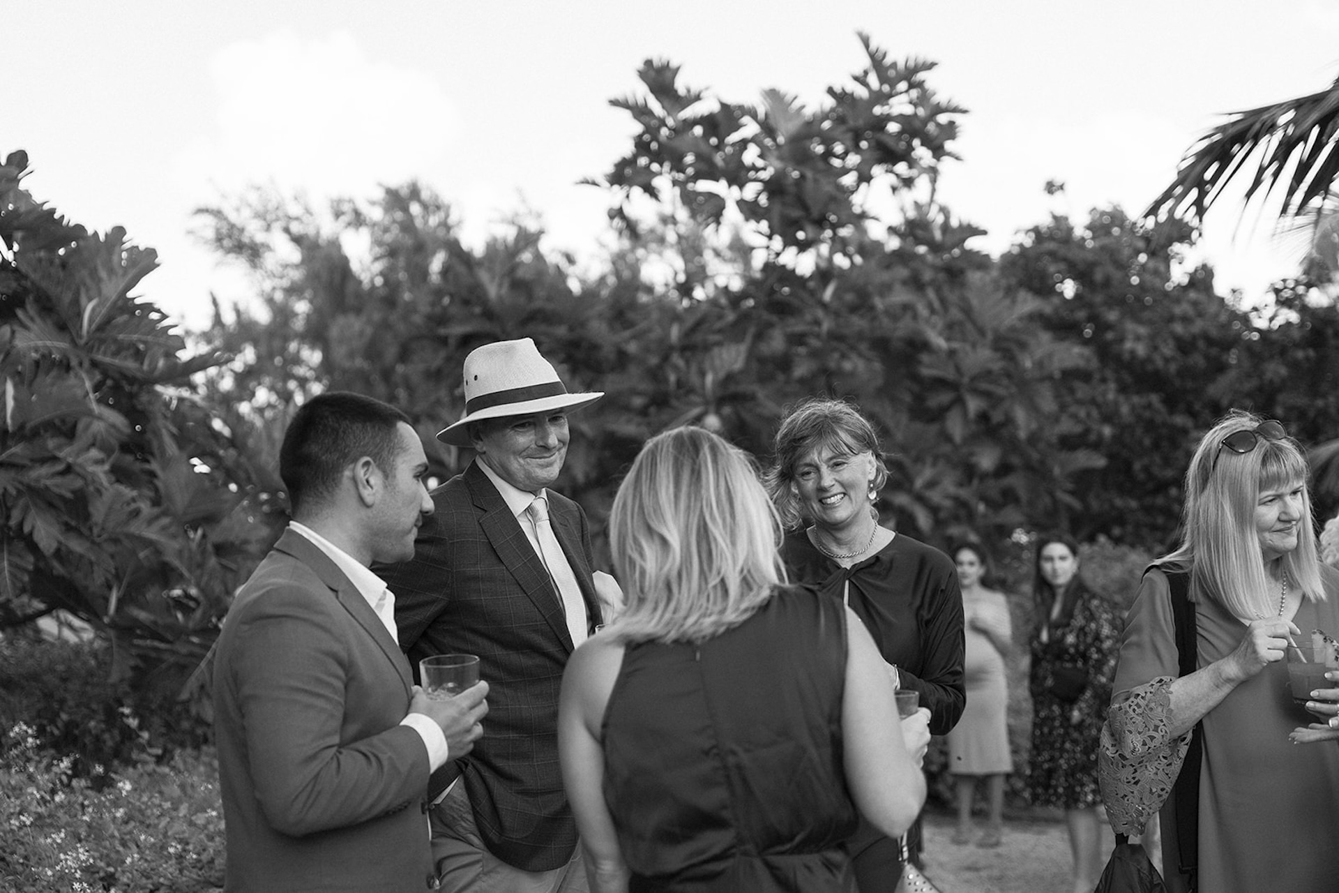 Black and white photo of wedding guests laughing and chatting during cocktail hour surrounded by tropical greenery.