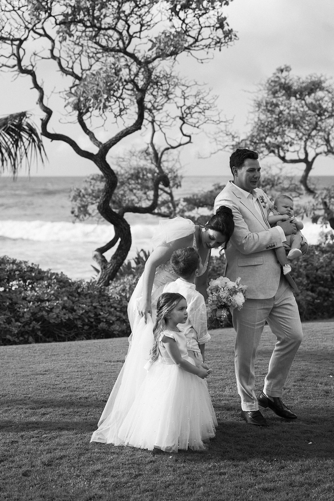 The bride and groom walk across the lawn with their children, the groom holding a baby while the flower girl leads ahead—a sweet family moment in the wedding weekend itinerary.