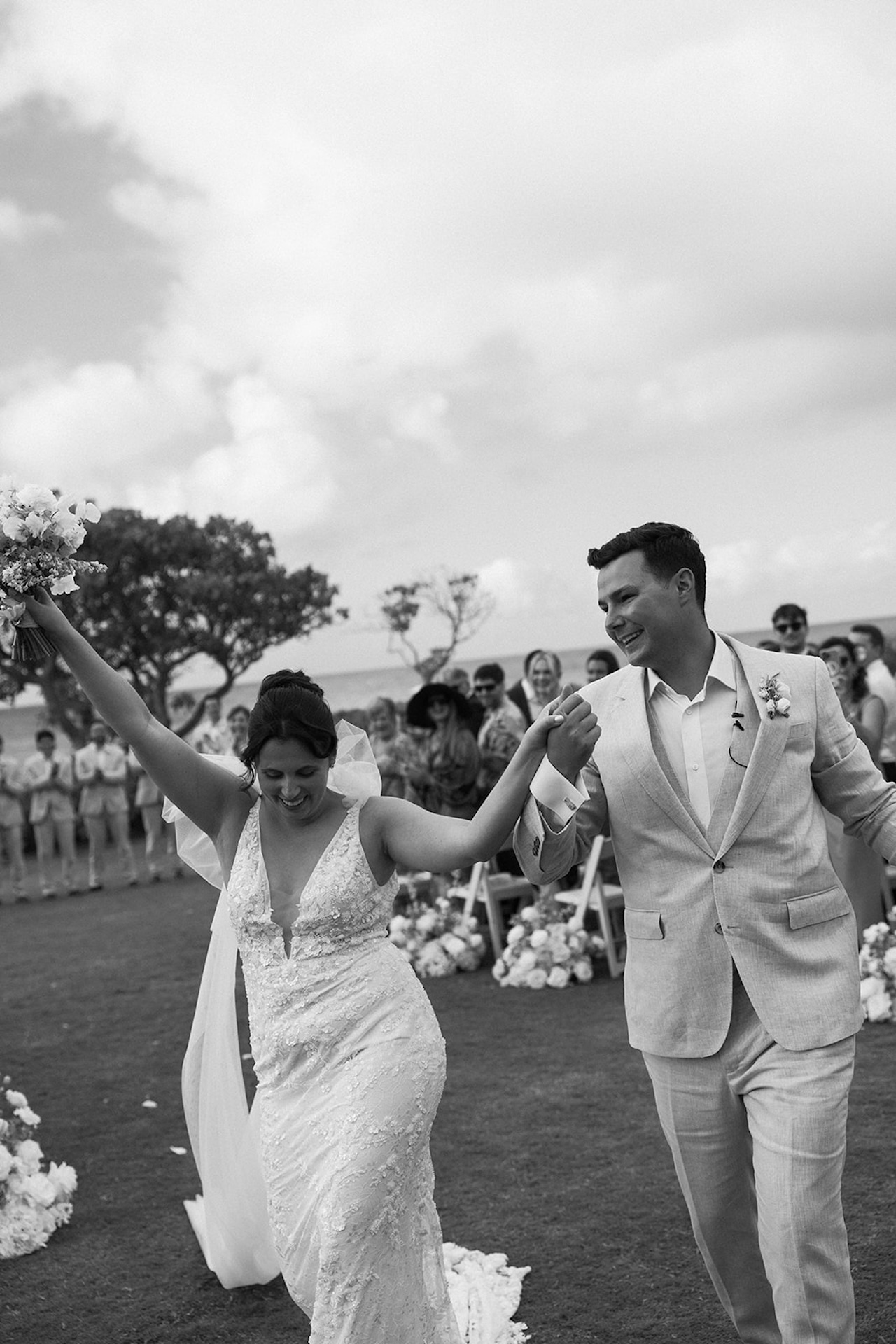 Black and white photo of the bride and groom joyfully walking back down the aisle, hands raised in celebration, marking the close of their wedding weekend itinerary ceremony.