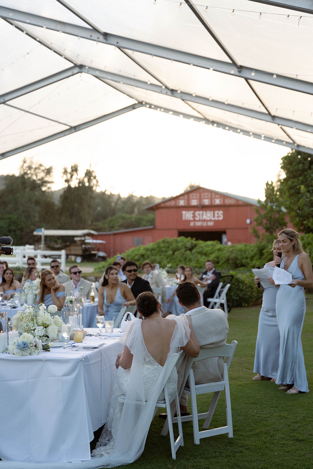 The bride and groom sit hand in hand as bridesmaids give speeches at the reception, laughter echoing across the tent, part of their wedding weekend itinerary.