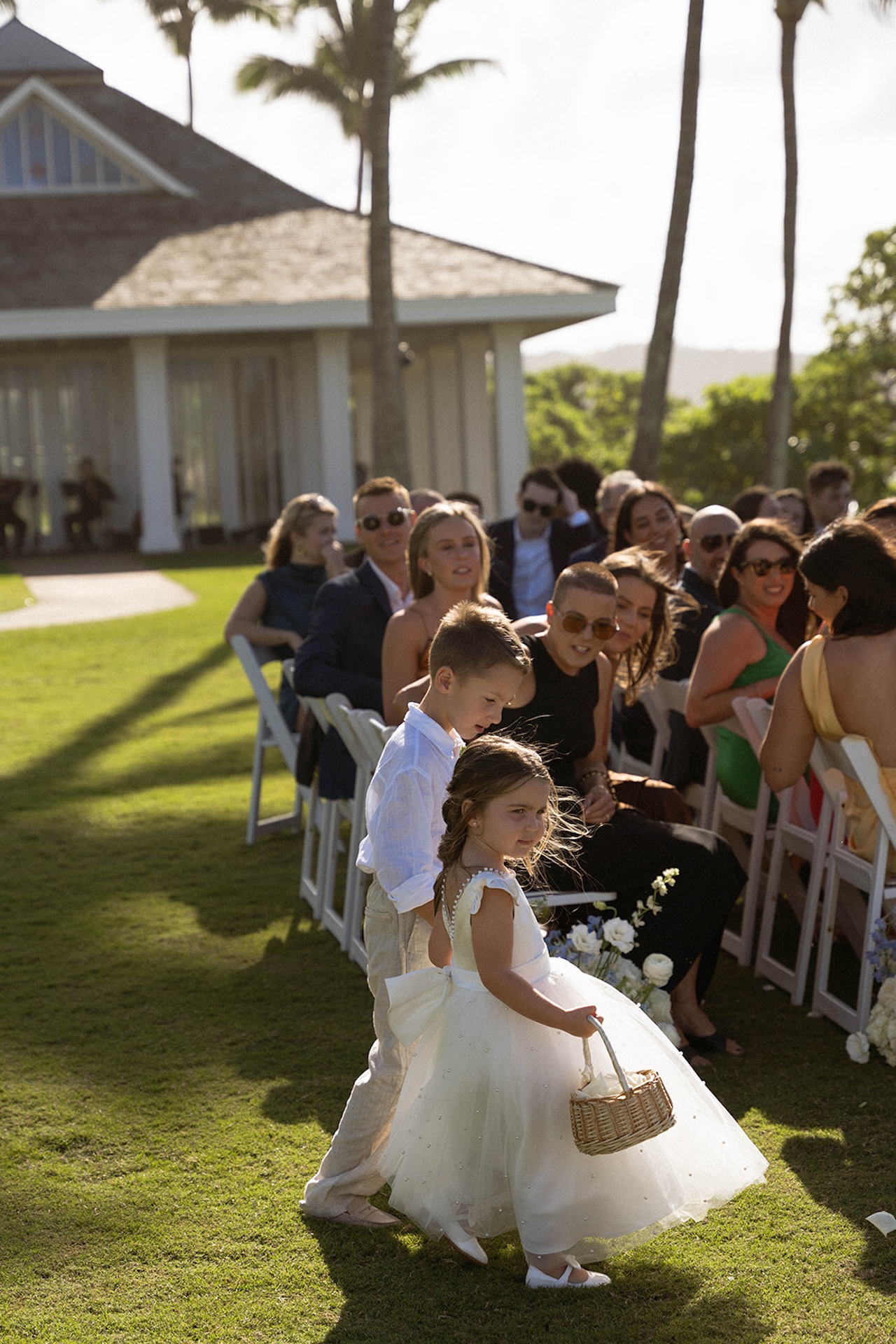 A flower girl and ring bearer walk hand in hand down the aisle, basket in hand, as wedding guests smile in the background.