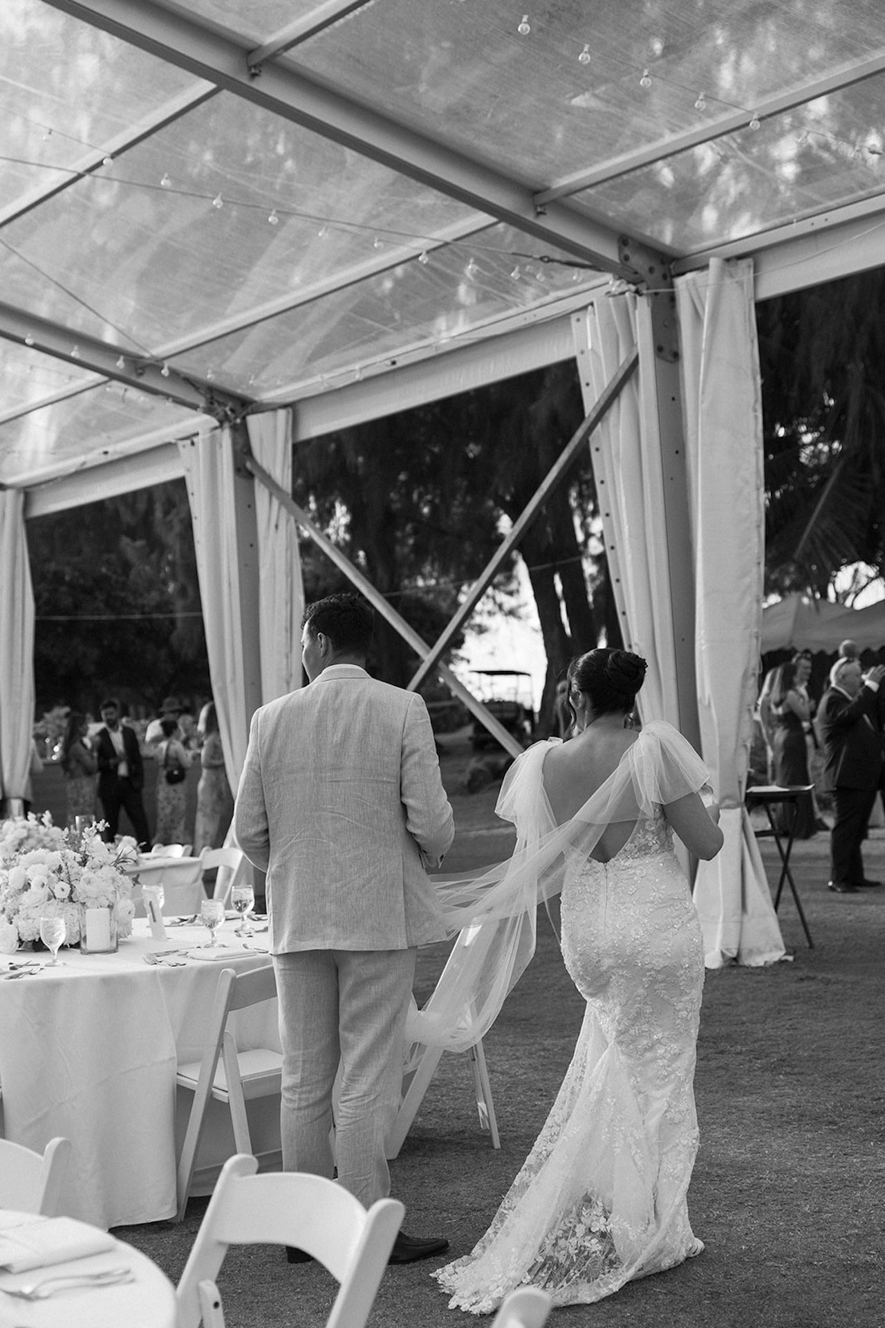 Black and white photo of the bride and groom entering the reception tent hand in hand, her long veil trailing behind her.