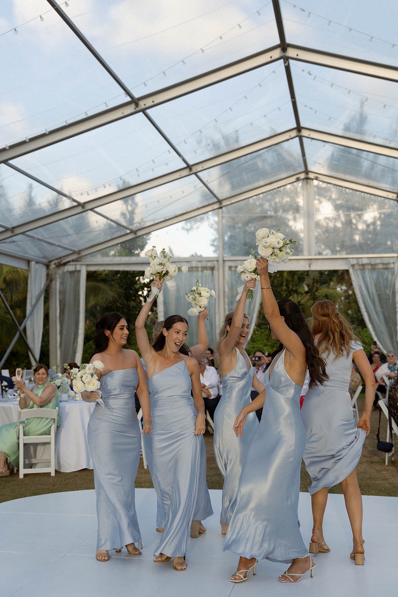 Bridesmaids in soft blue gowns lift their bouquets high on the dance floor, celebrating together during the evening reception, part of the couple’s wedding weekend itinerary.