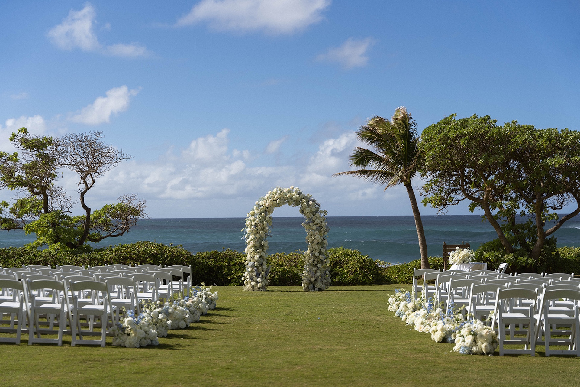 Oceanfront ceremony setup featuring a circular floral arch framed by palm trees and rows of white chairs, waiting for guests to arrive.