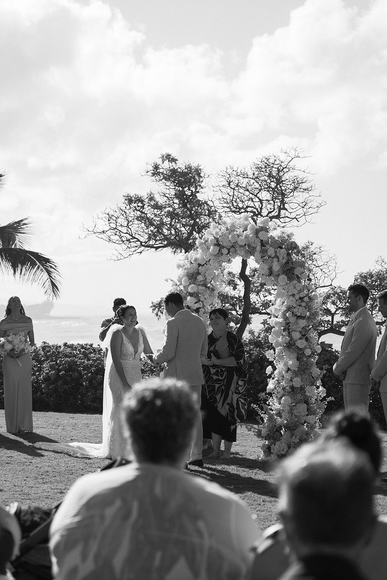 The bride and groom exchange vows beneath a floral arch as sunlight streams across the ceremony, guests watching intently.