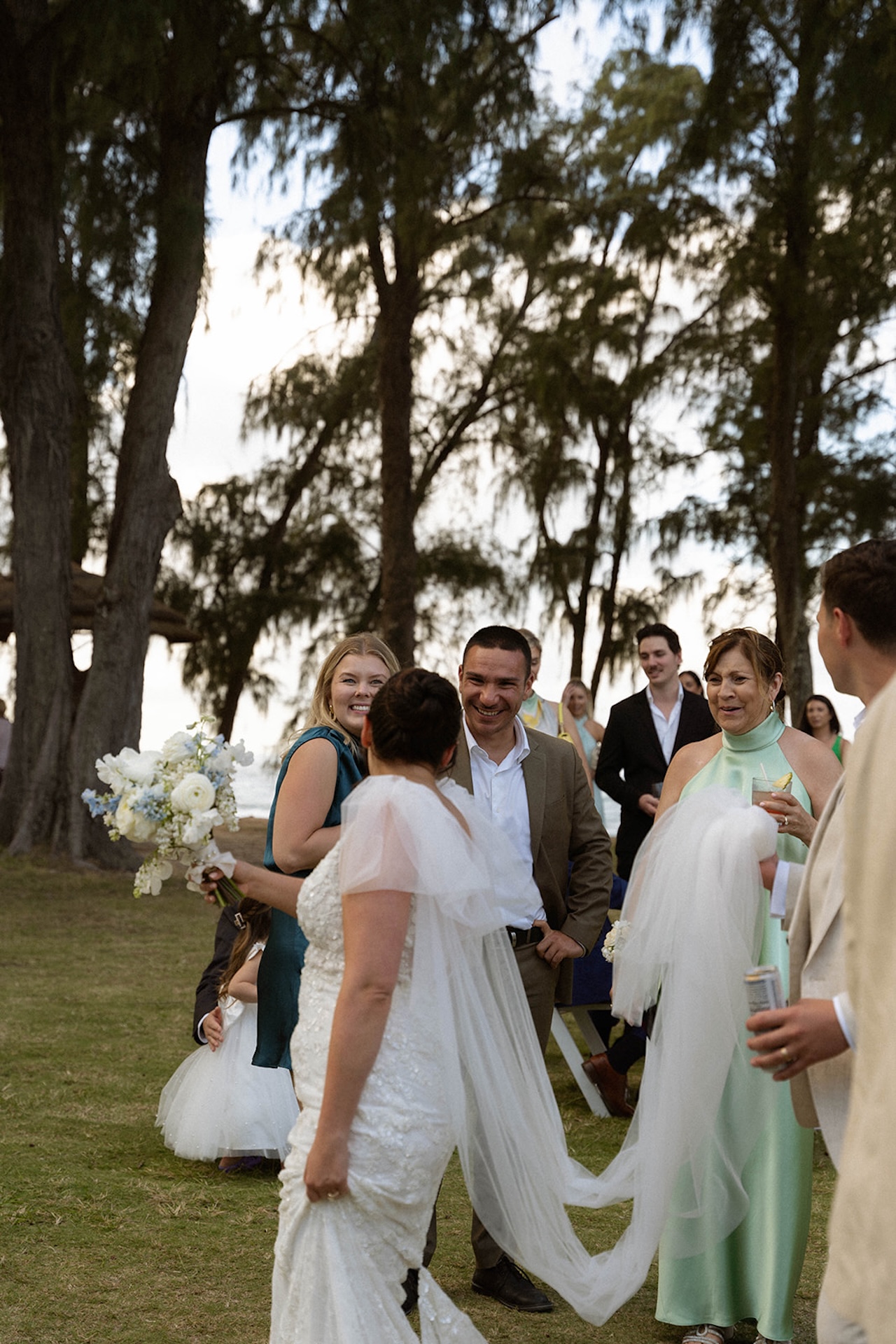 The bride greets smiling guests outside the reception tent, her bouquet in hand and veil trailing behind her, adding warmth to the wedding weekend itinerary.
