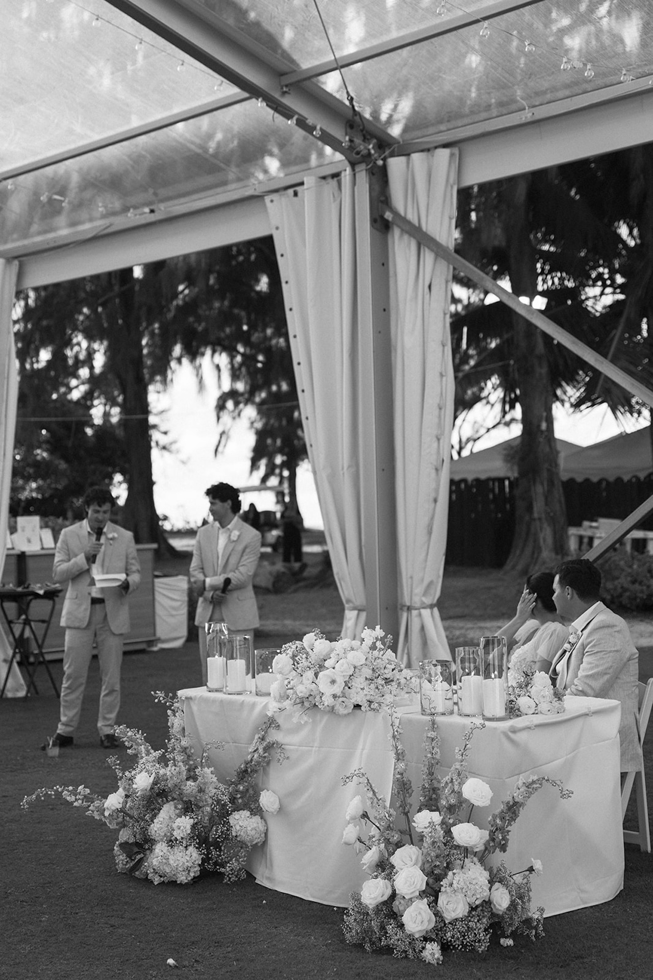 The bride and groom sit at a sweetheart table surrounded by abundant florals as groomsmen give speeches during the reception.