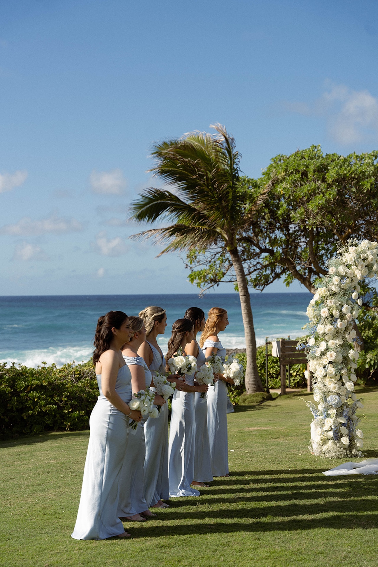 Bridesmaids in light blue dresses line up by the oceanfront ceremony arch, bouquets of white flowers in hand, ready for the next part of the wedding weekend itinerary.