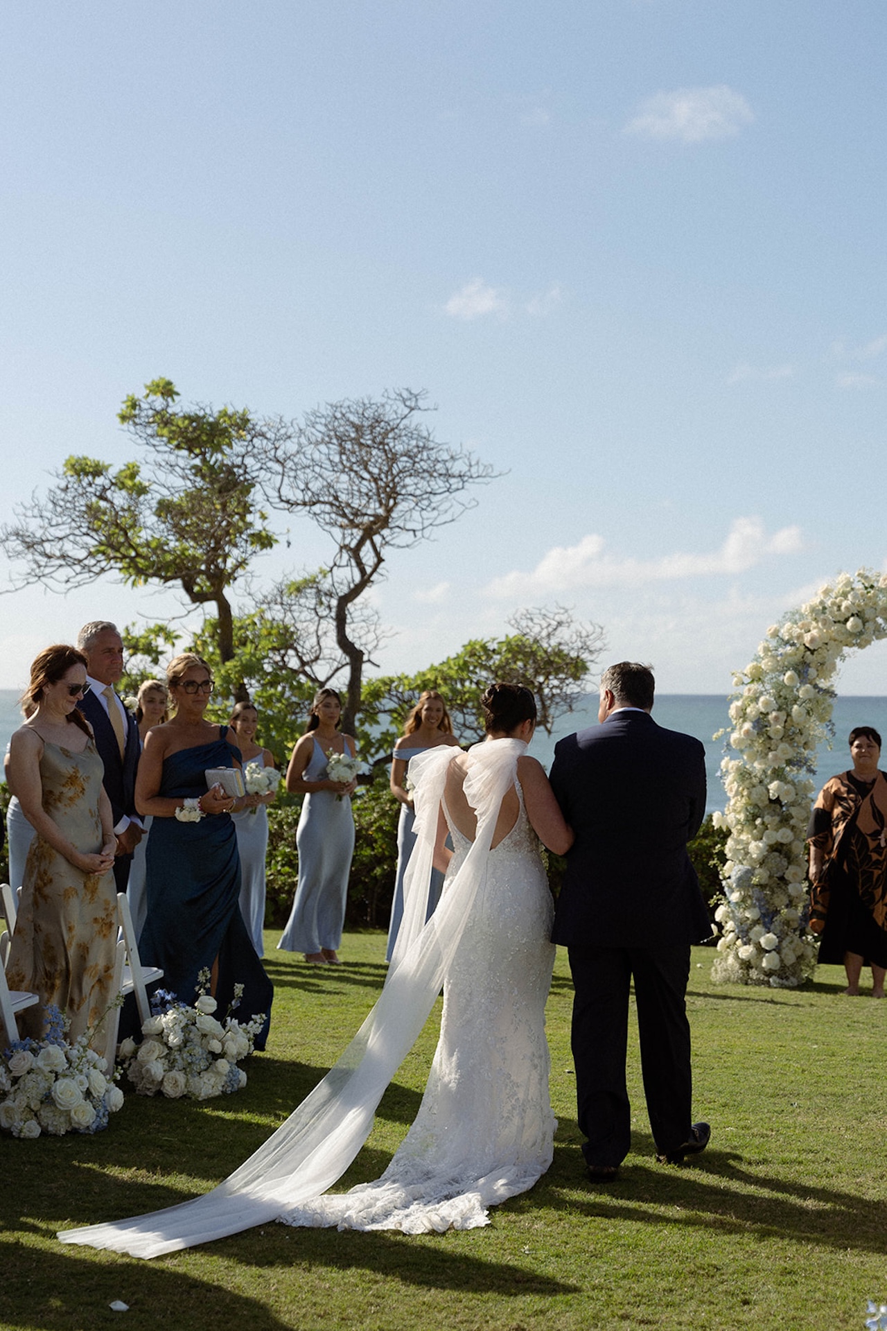 The bride is escorted down the aisle toward a floral arch overlooking the ocean, with bridesmaids and family waiting ahead.