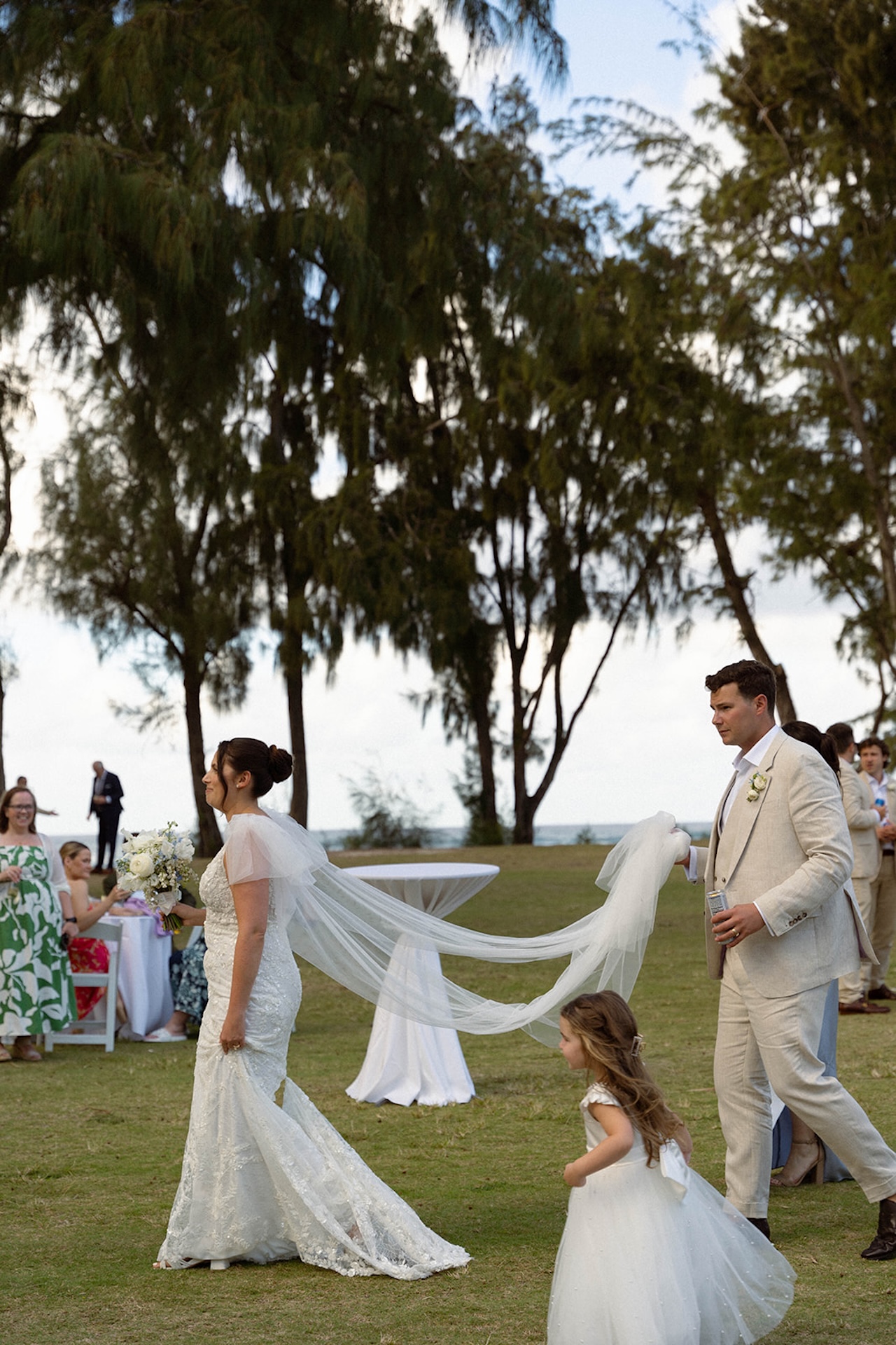 The bride walks across the lawn with her groom and flower girl, her veil flowing behind her, a sweet moment that highlights the start of their wedding weekend itinerary.