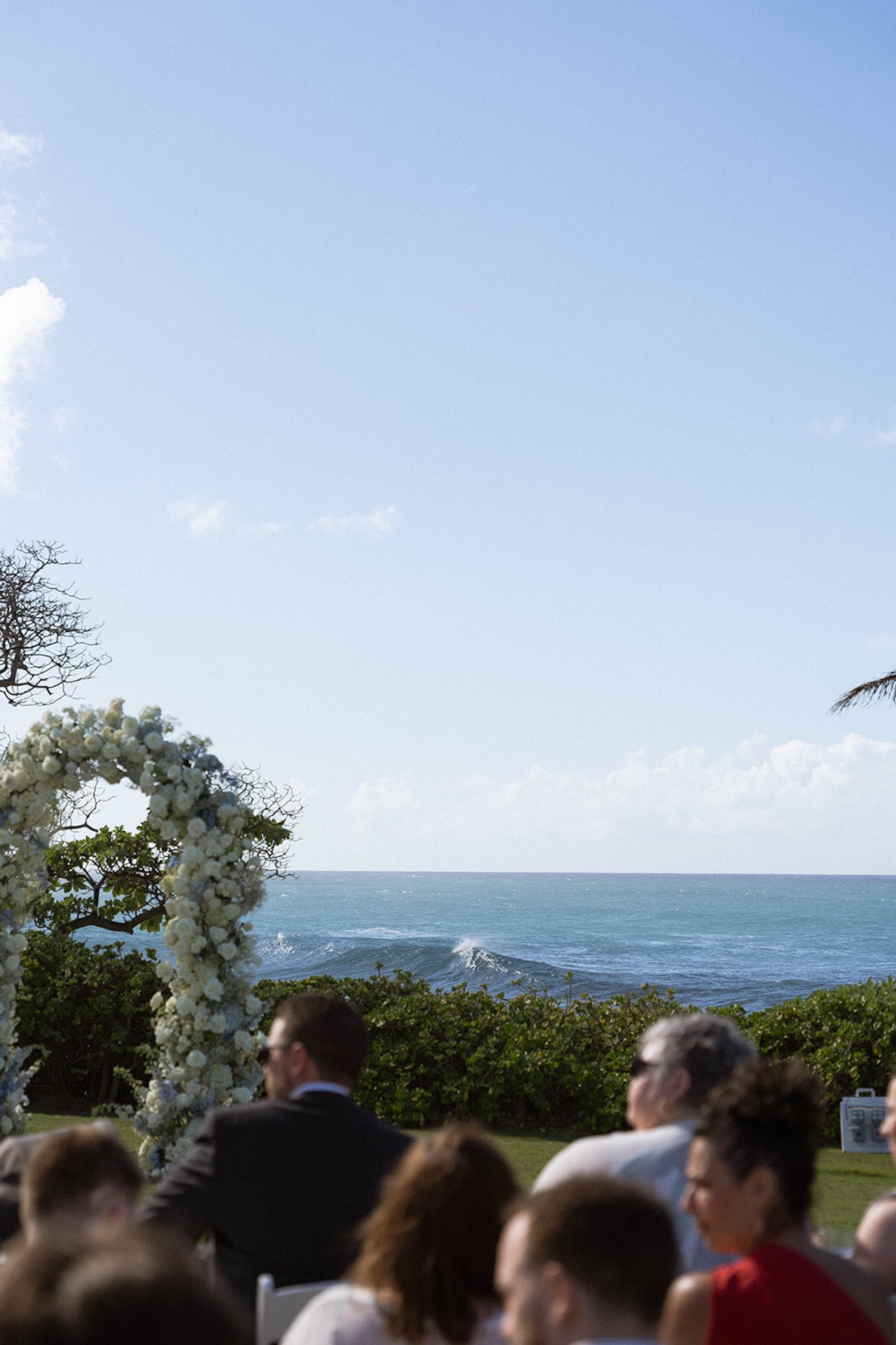 Guests look toward the ocean during the ceremony, framed by a large floral arch, waves rolling in the background—a serene moment in the wedding weekend itinerary.