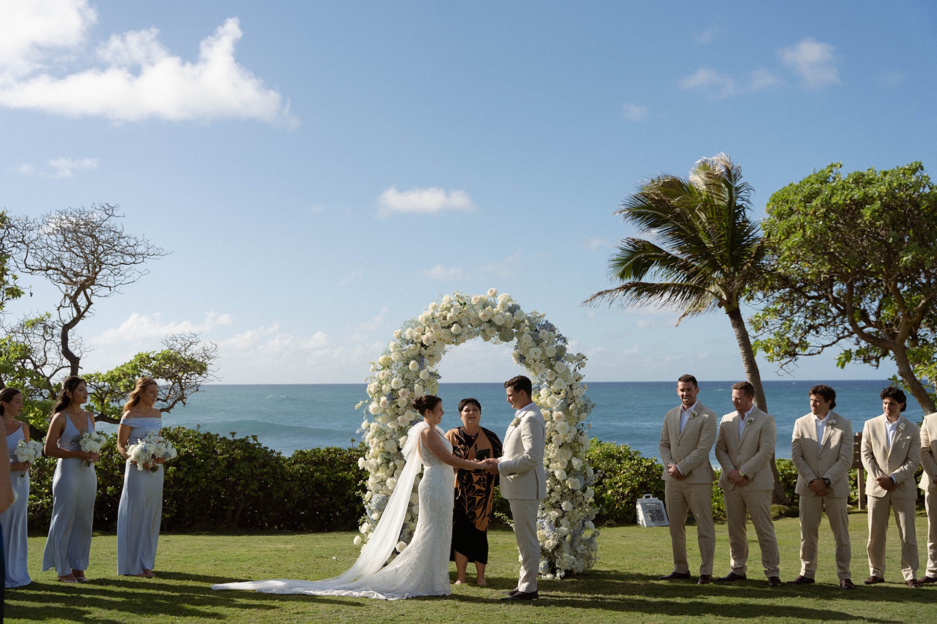 The bride and groom stand before the officiant under a large circular floral arch, exchanging vows with the ocean behind them.