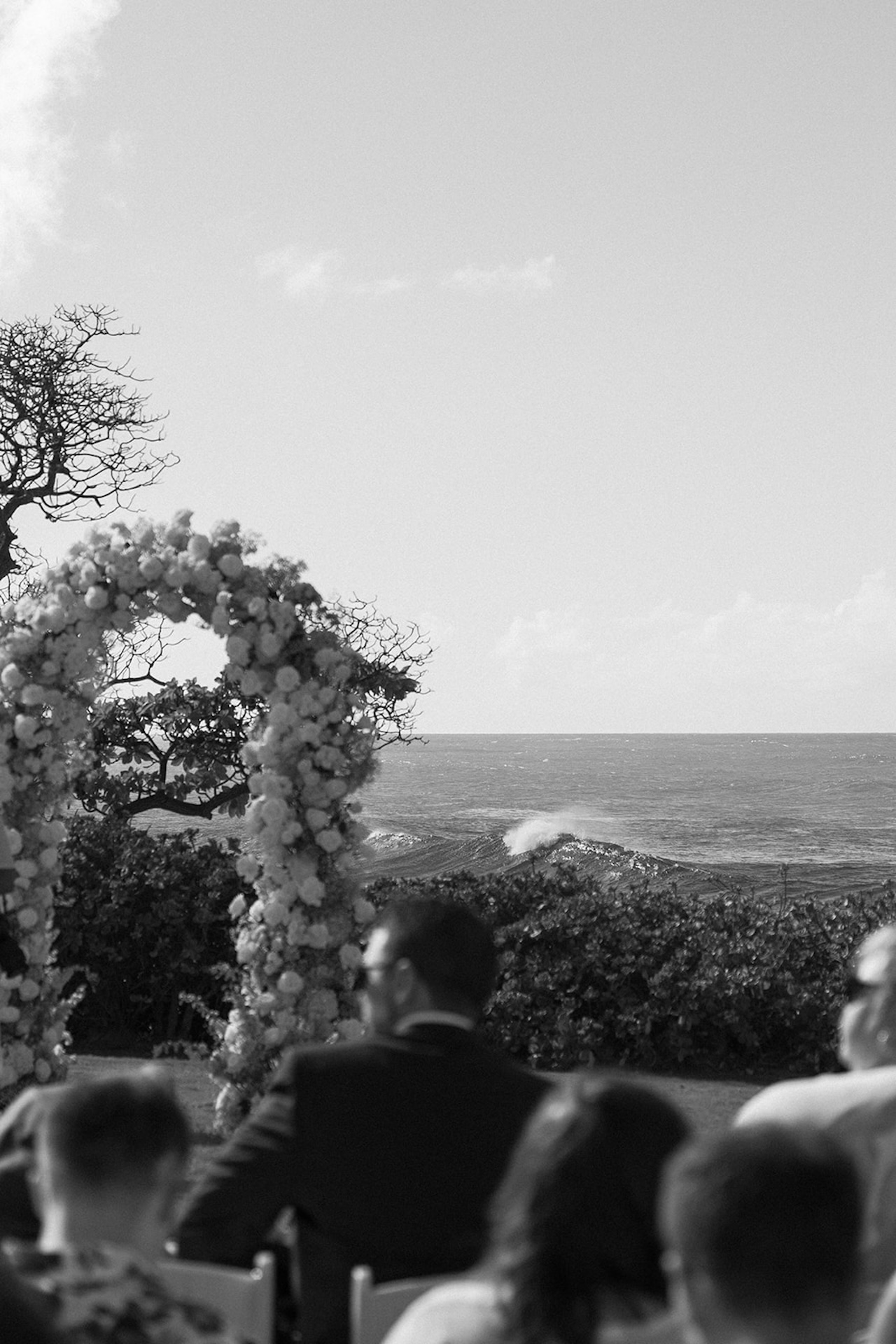 Black and white photo of guests seated at the ceremony, with the floral arch framing waves crashing on the ocean beyond.