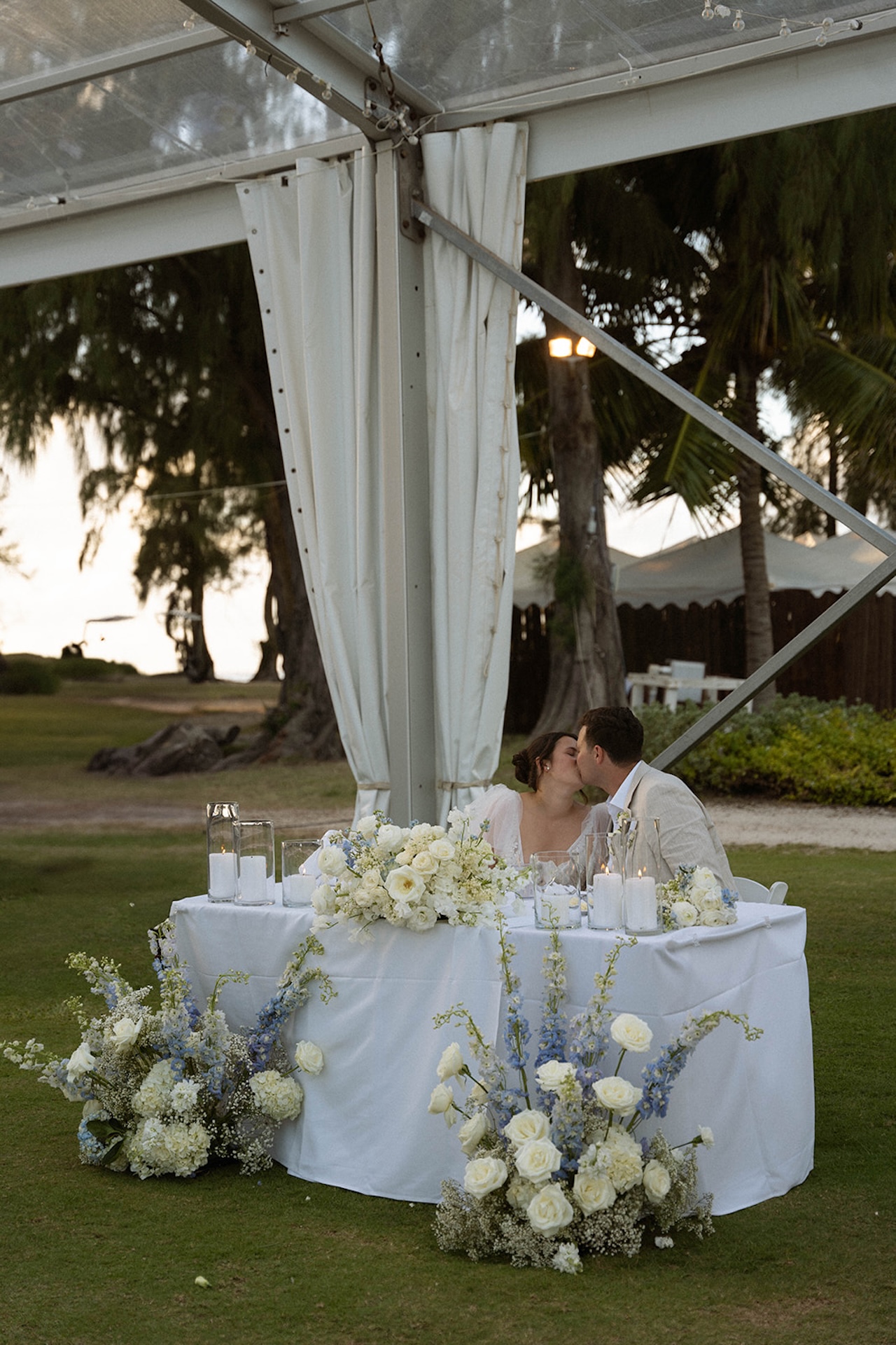 The bride and groom share a kiss at their sweetheart table, decorated with white and blue florals, a romantic reception highlight in the wedding weekend itinerary.