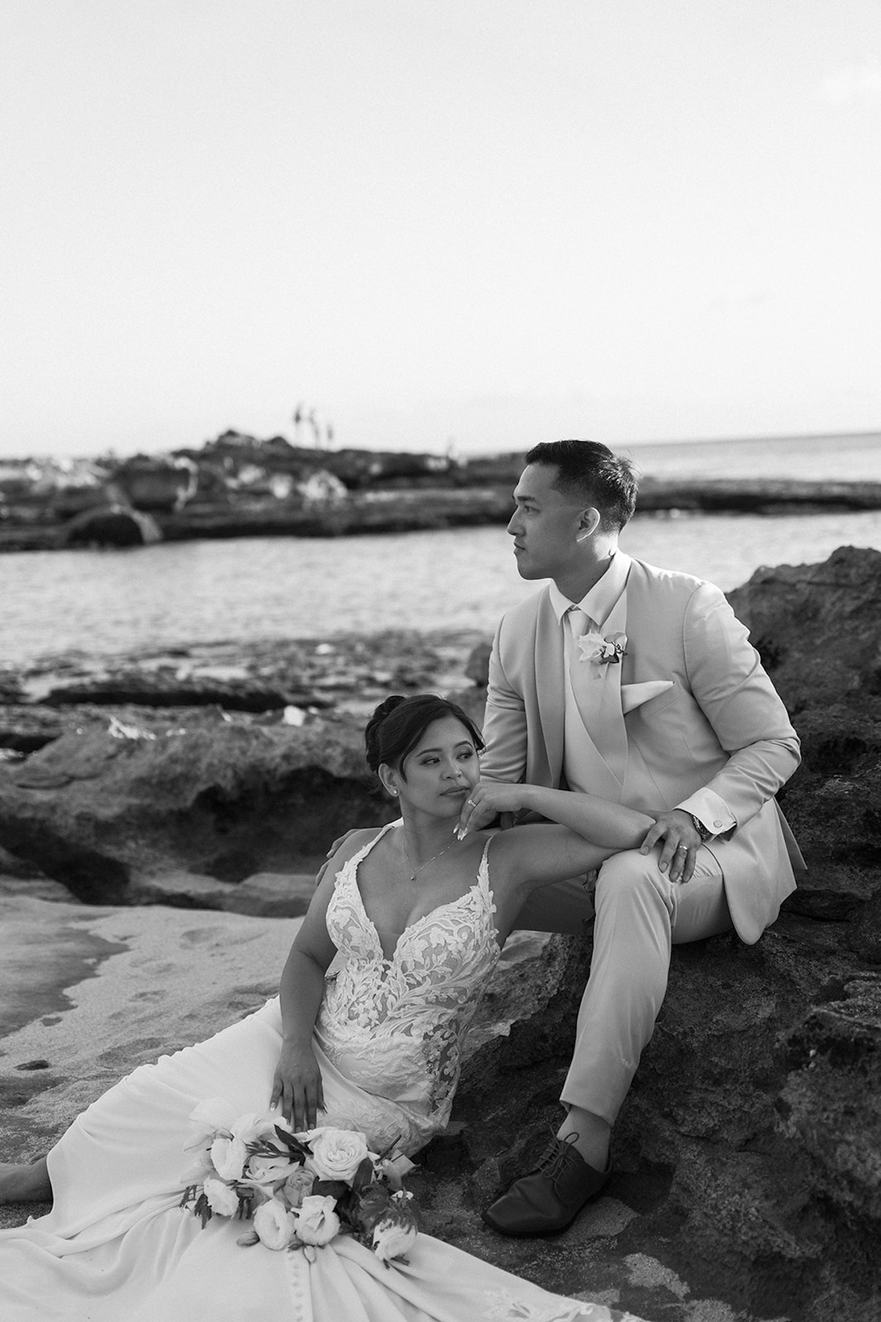 A black-and-white photo of a newlywed couple posing on the rocks by the ocean, capturing timeless coastal romance.