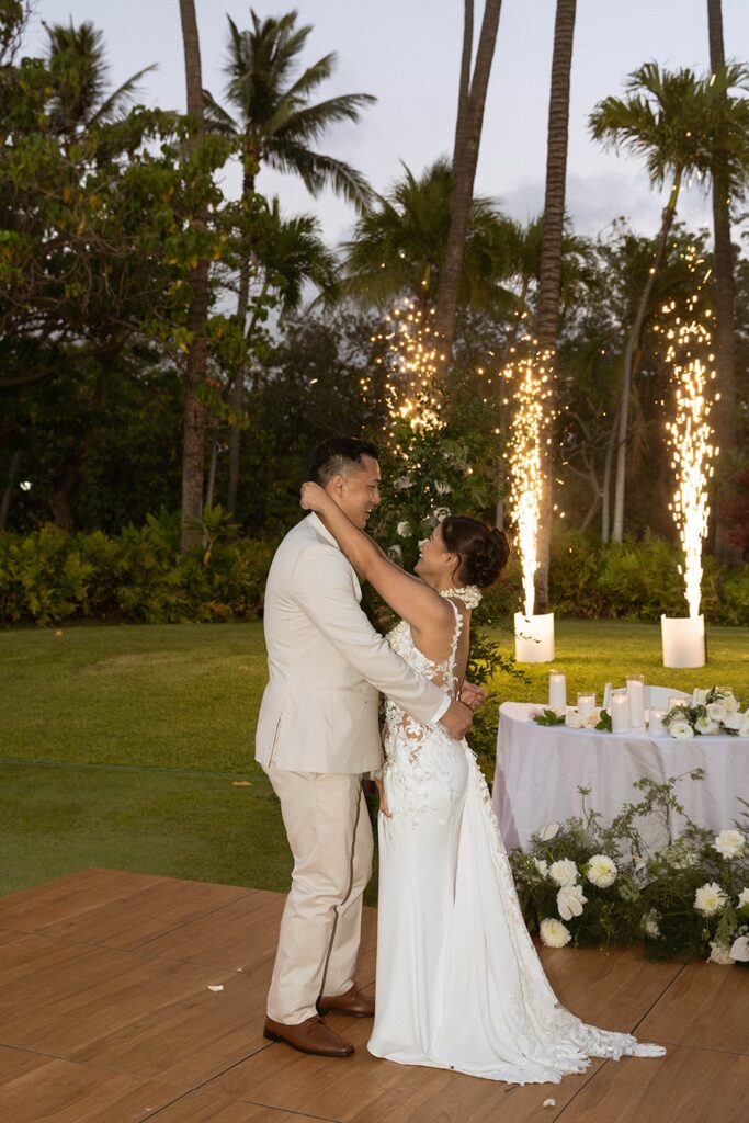 The couple embraces during their first dance beneath glowing sparklers, framed by tropical greenery and palm trees swaying in the evening breeze.