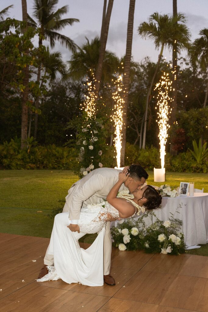 The groom dips the bride into a romantic kiss during their first dance, surrounded by sparklers and tropical palm trees at their outdoor Hawaii reception.