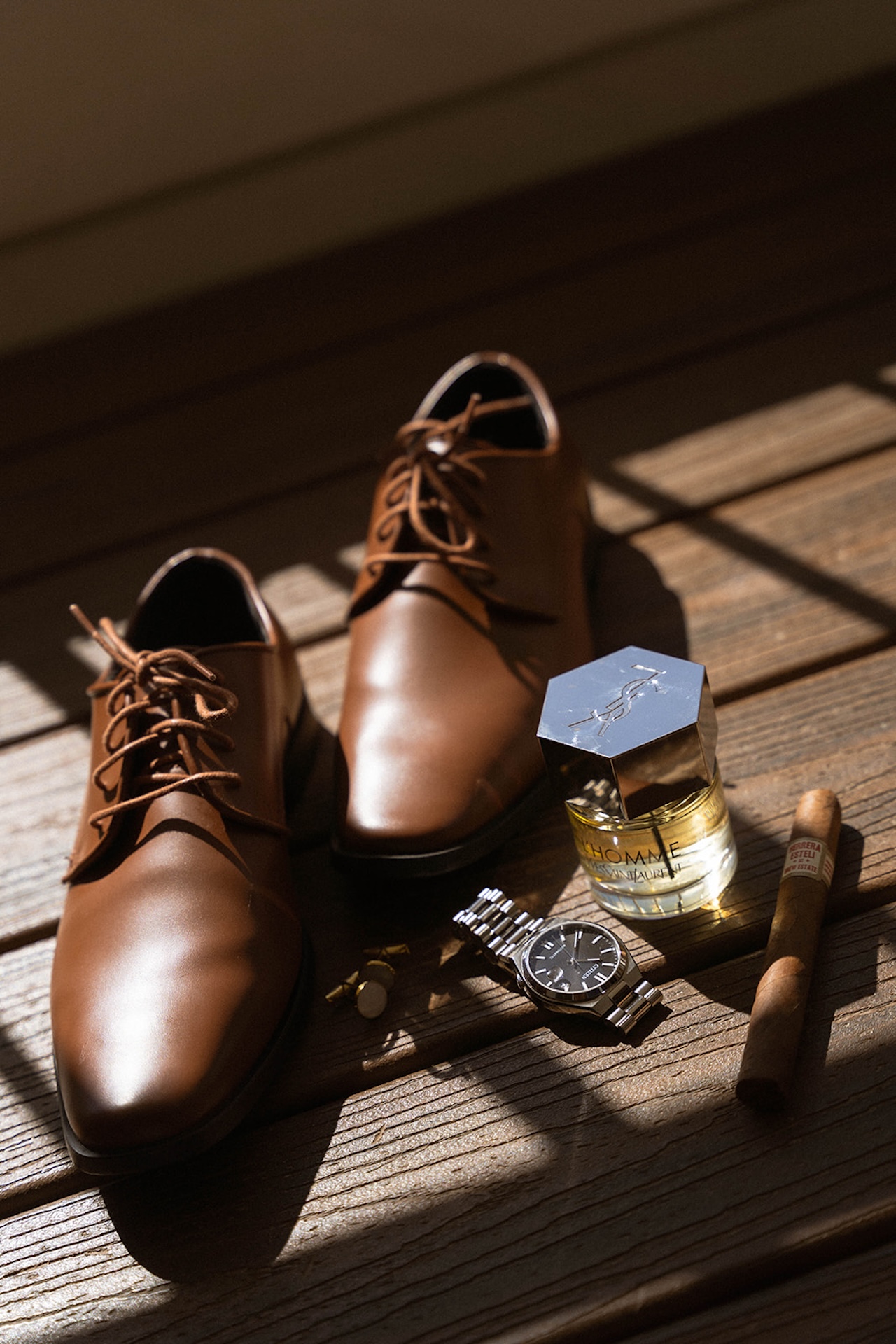 Groom’s flatlay details on wooden flooring, brown leather shoes, YSL cologne, silver watch, cigar, and cufflinks, timeless accessories for any Destination wedding checklist.