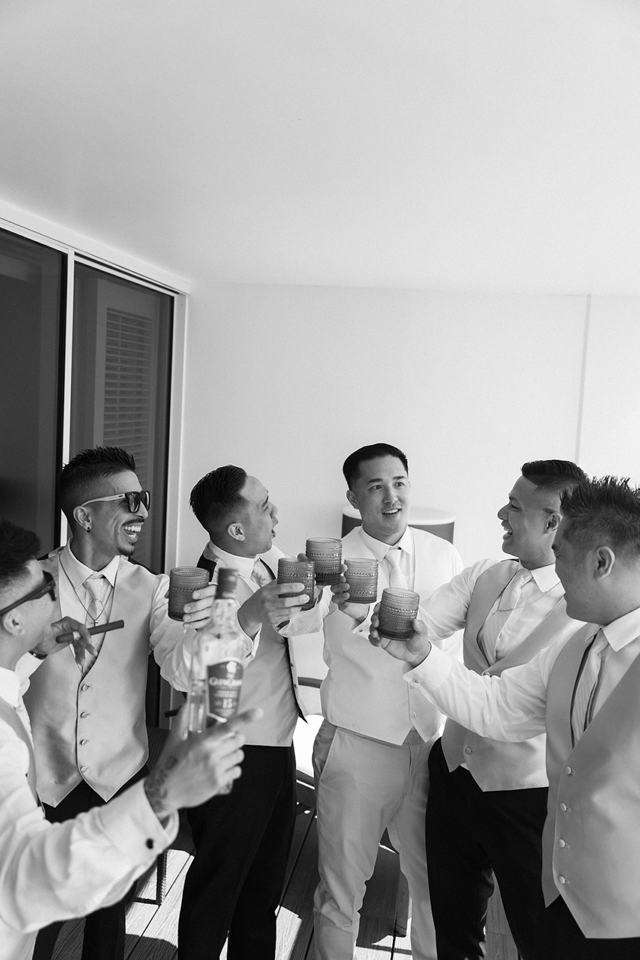 Black-and-white photo of the groom and his groomsmen toasting with drinks and laughing together before the ceremony.