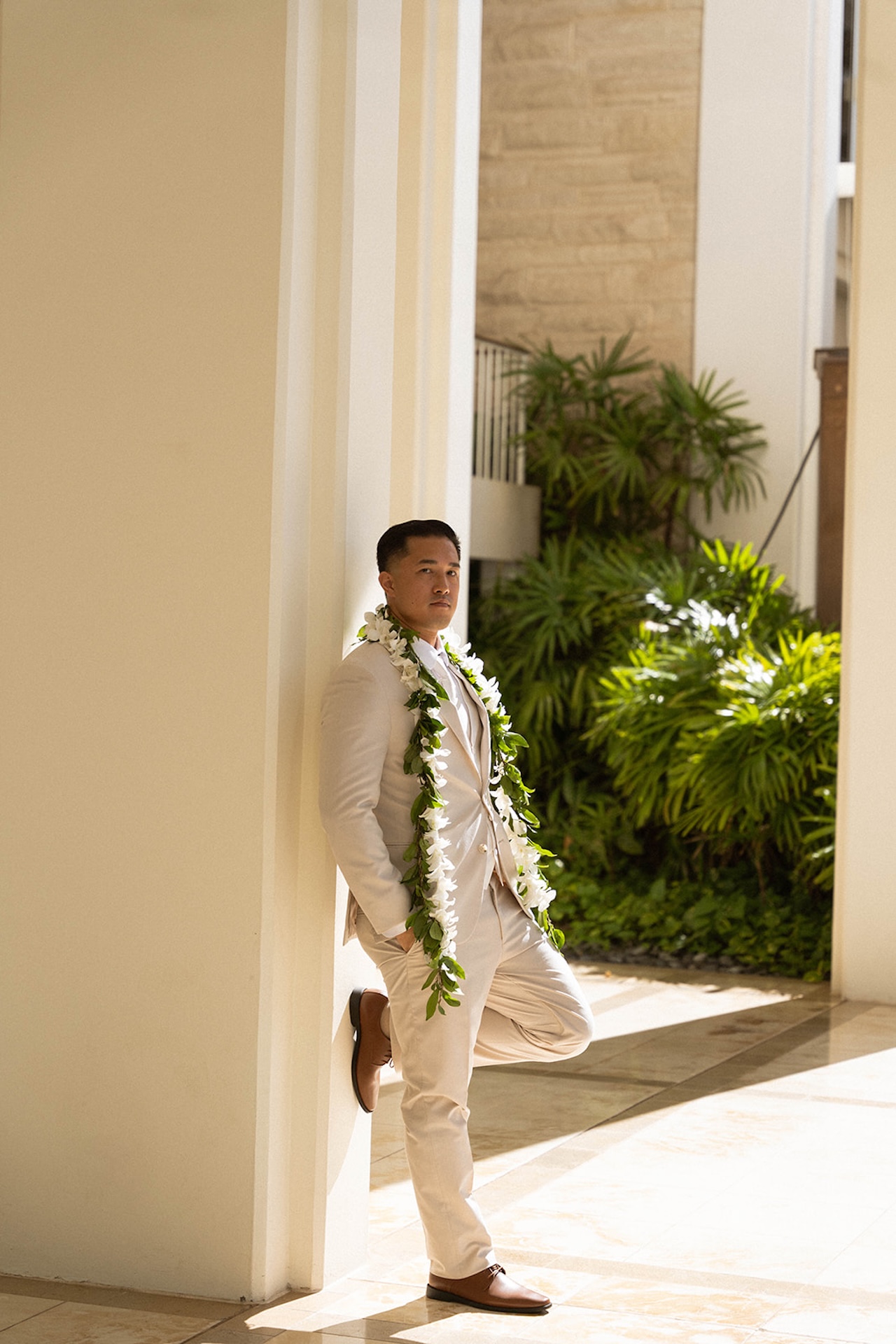 The groom leans casually against a sunlit column in a beige suit adorned with a white floral lei, a relaxed yet elegant look fitting for a Destination wedding checklist celebration.