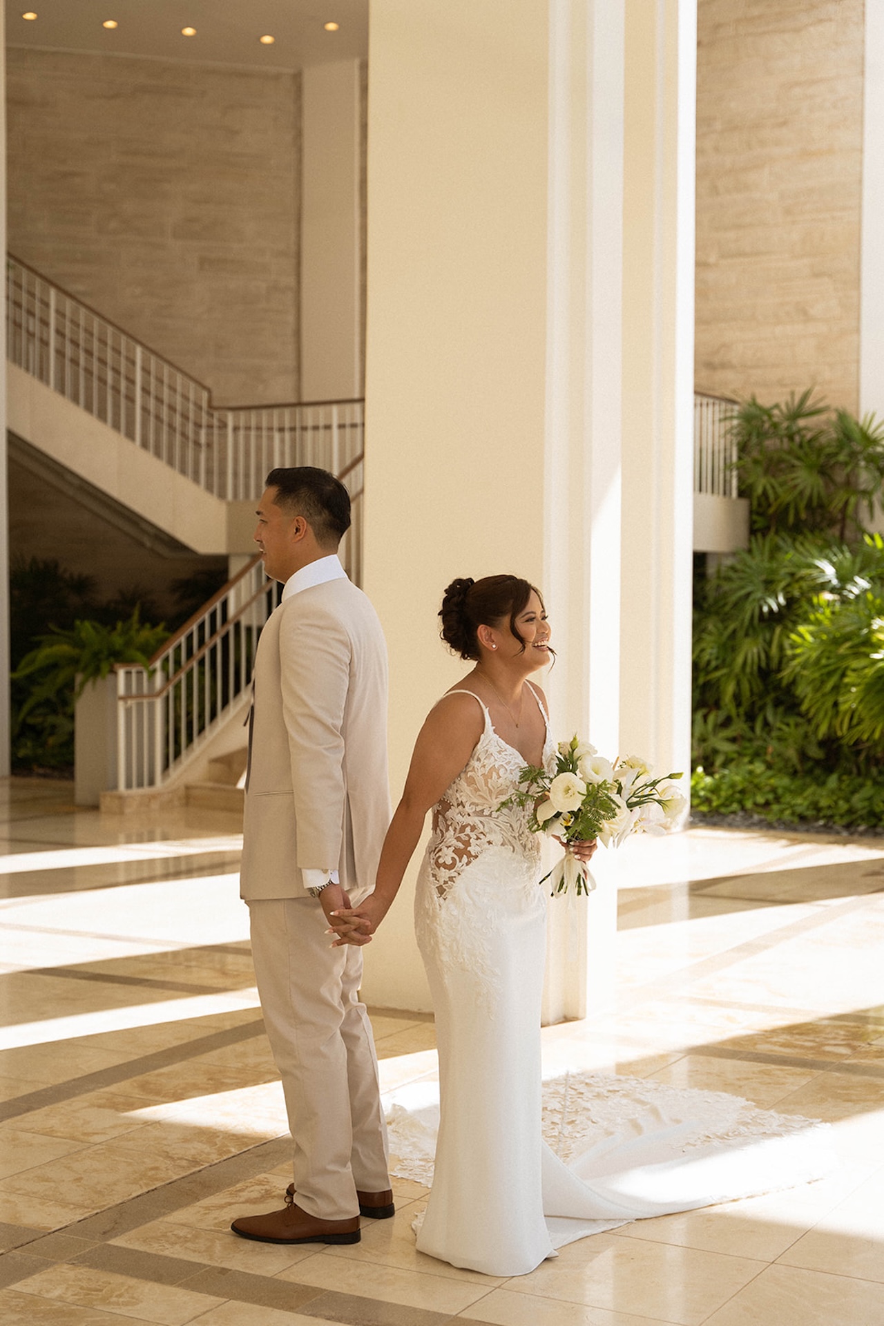 The couple stands back-to-back holding hands during their first look moment inside a bright, modern venue filled with natural light.