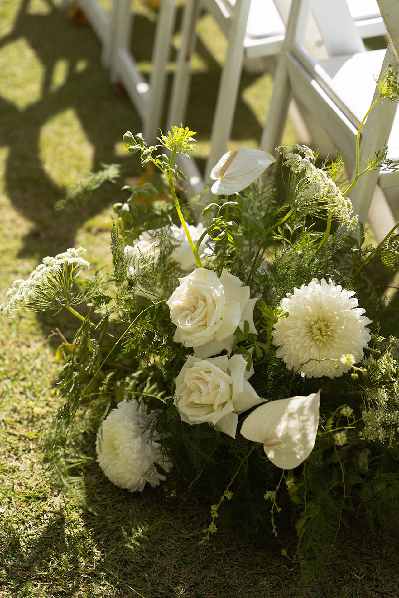 Close-up of white floral arrangements with roses, calla lilies, and greenery lining the ceremony aisle, softly lit by afternoon sun.