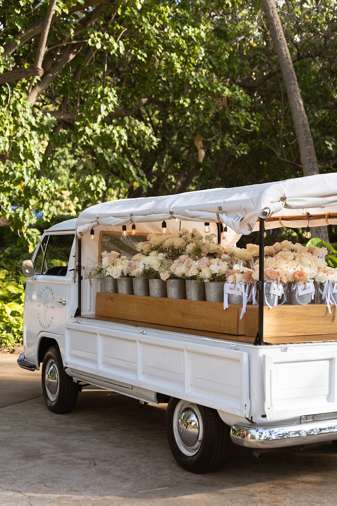 White vintage truck adorned with soft blooms and labeled table arrangements parked beneath tropical trees, a stylish floral setup for an island wedding.