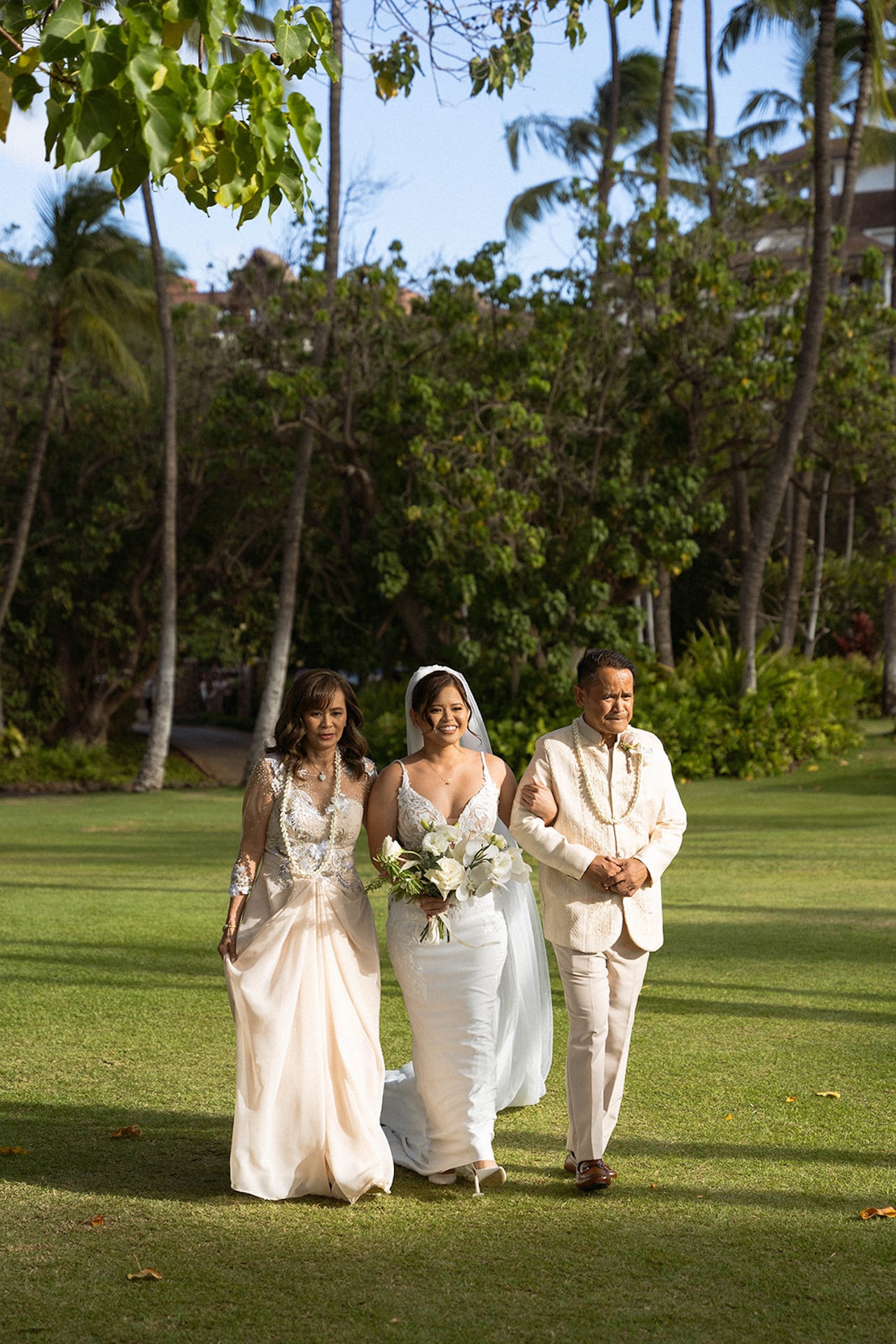The bride walking arm-in-arm with her parents down the lush green aisle, sunlight streaming through the trees.