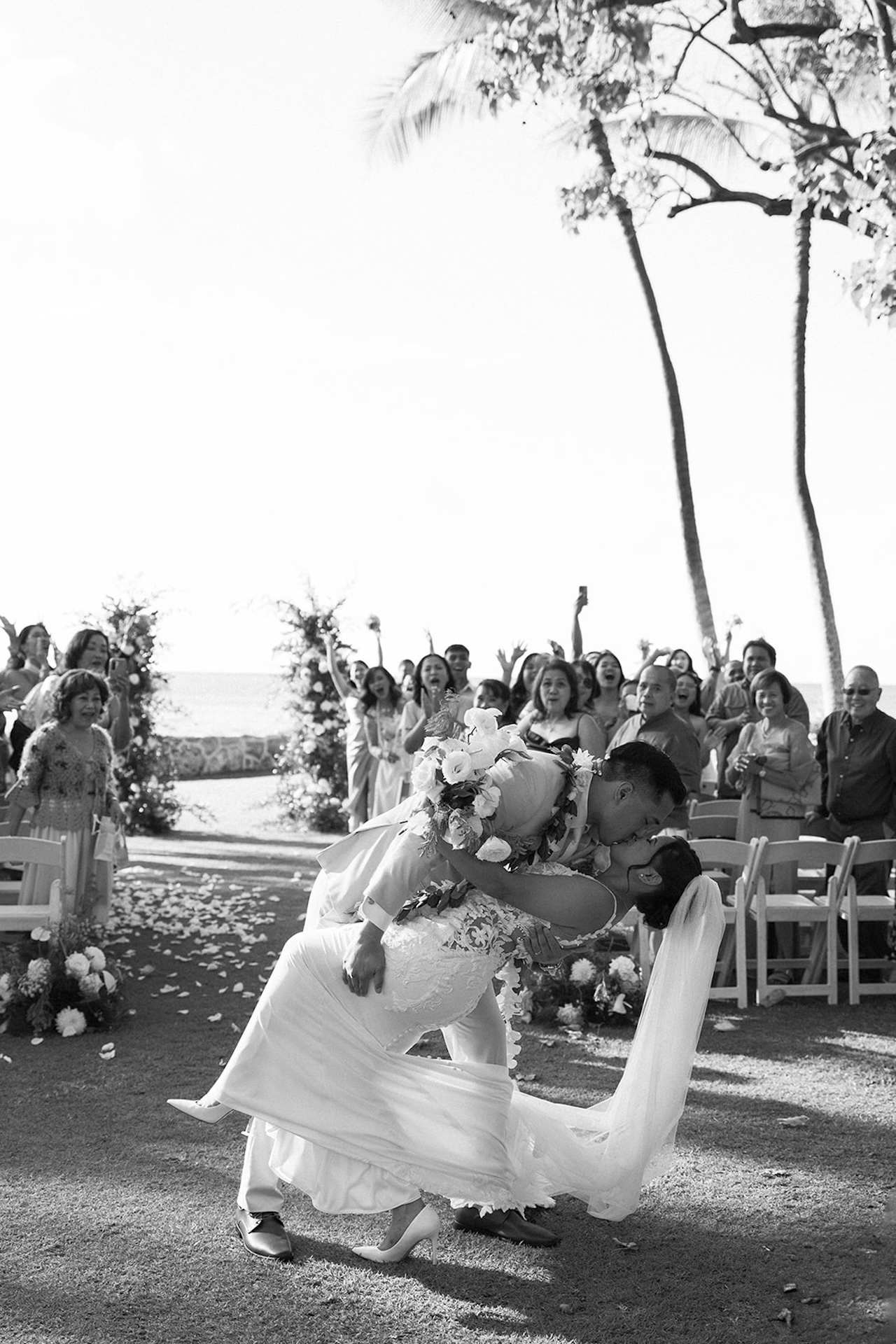 Black-and-white photo of the groom dipping the bride into a kiss as guests cheer behind them, framed by palm trees and ocean views.