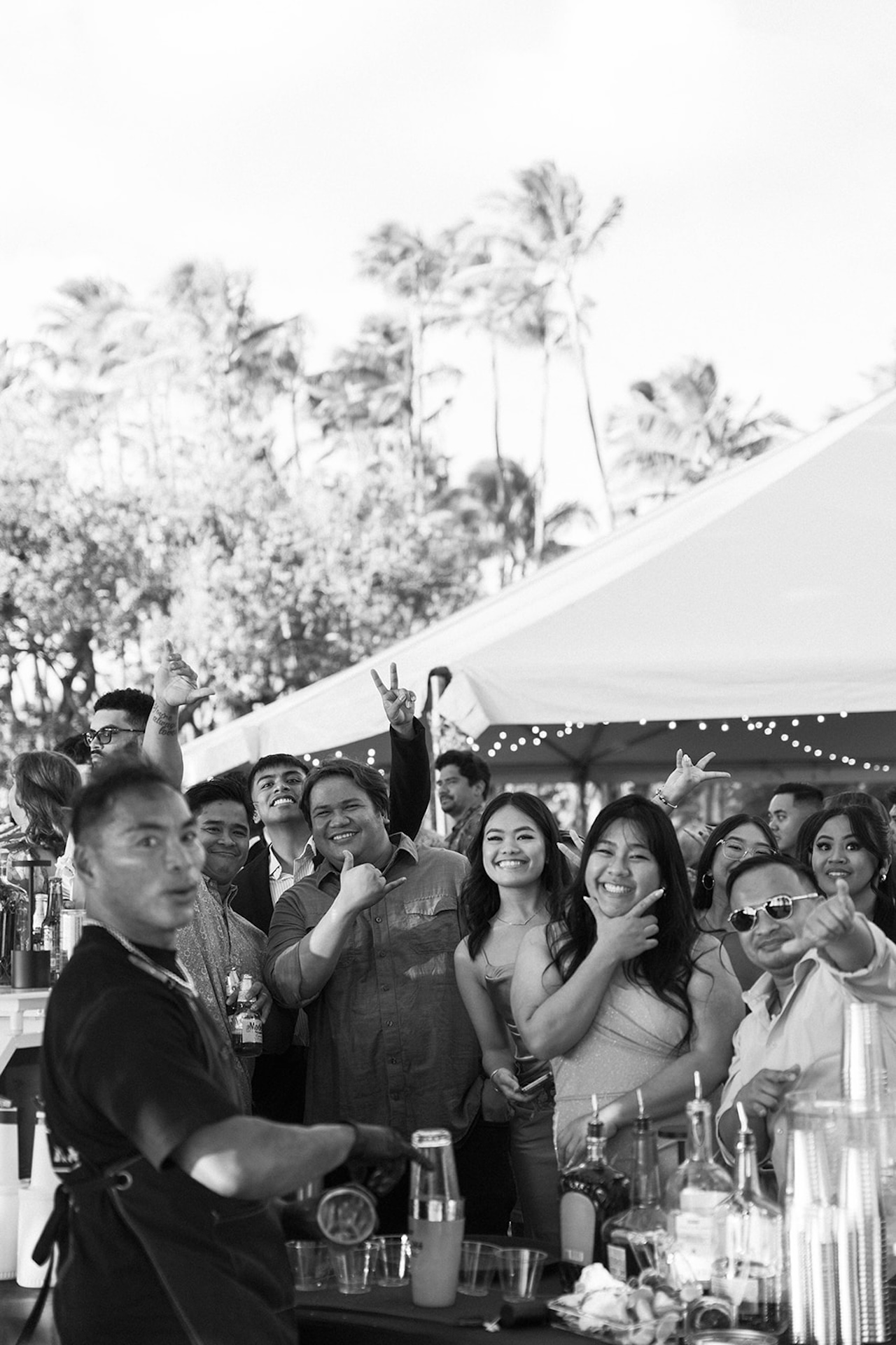 A lively group of guests gathered at the bar, laughing and cheering during cocktail hour under tent lights and palm trees.