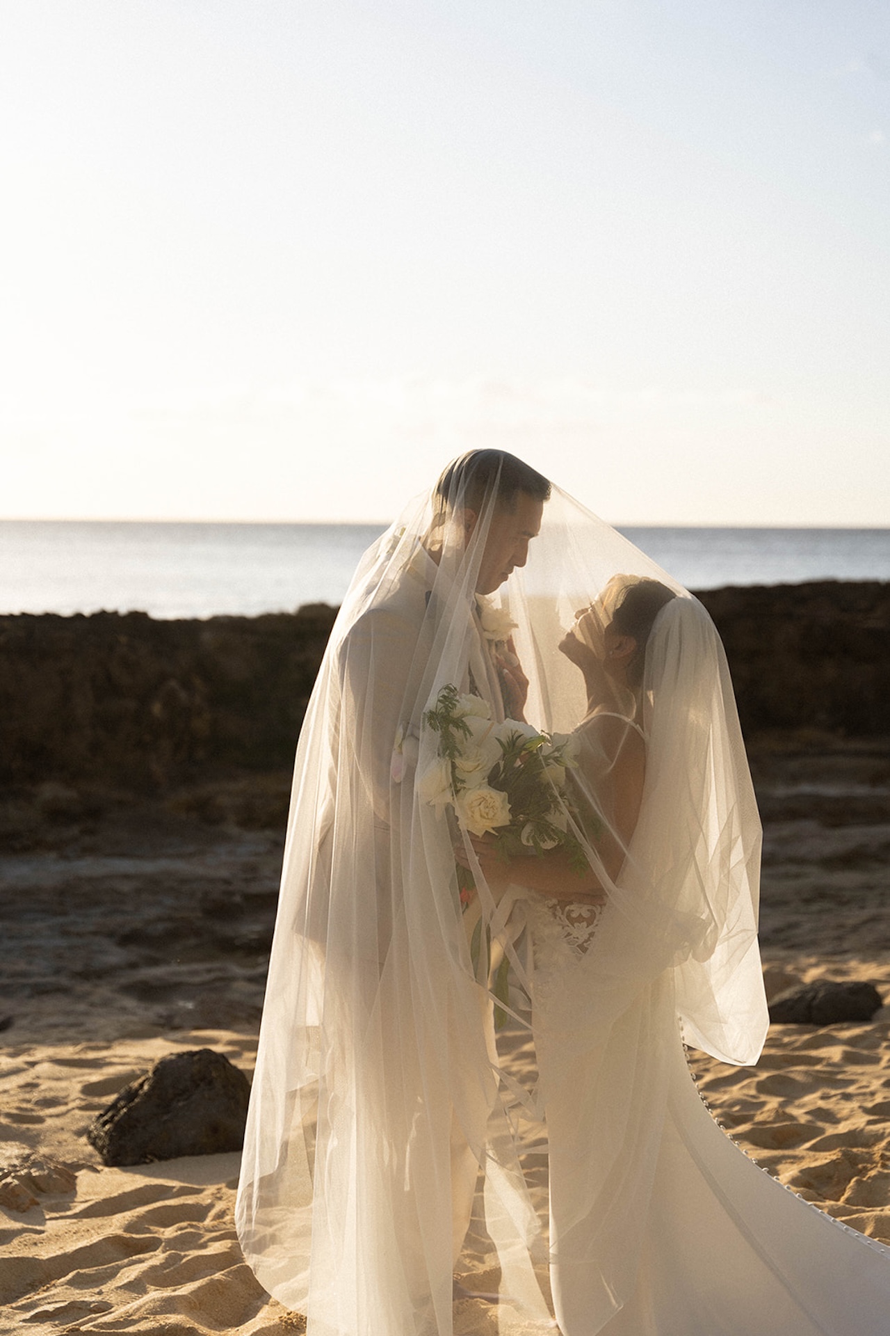 Couple under the bride’s veil at sunset, sharing an intimate moment on the sandy shore, the perfect ending to a Destination wedding checklist love story