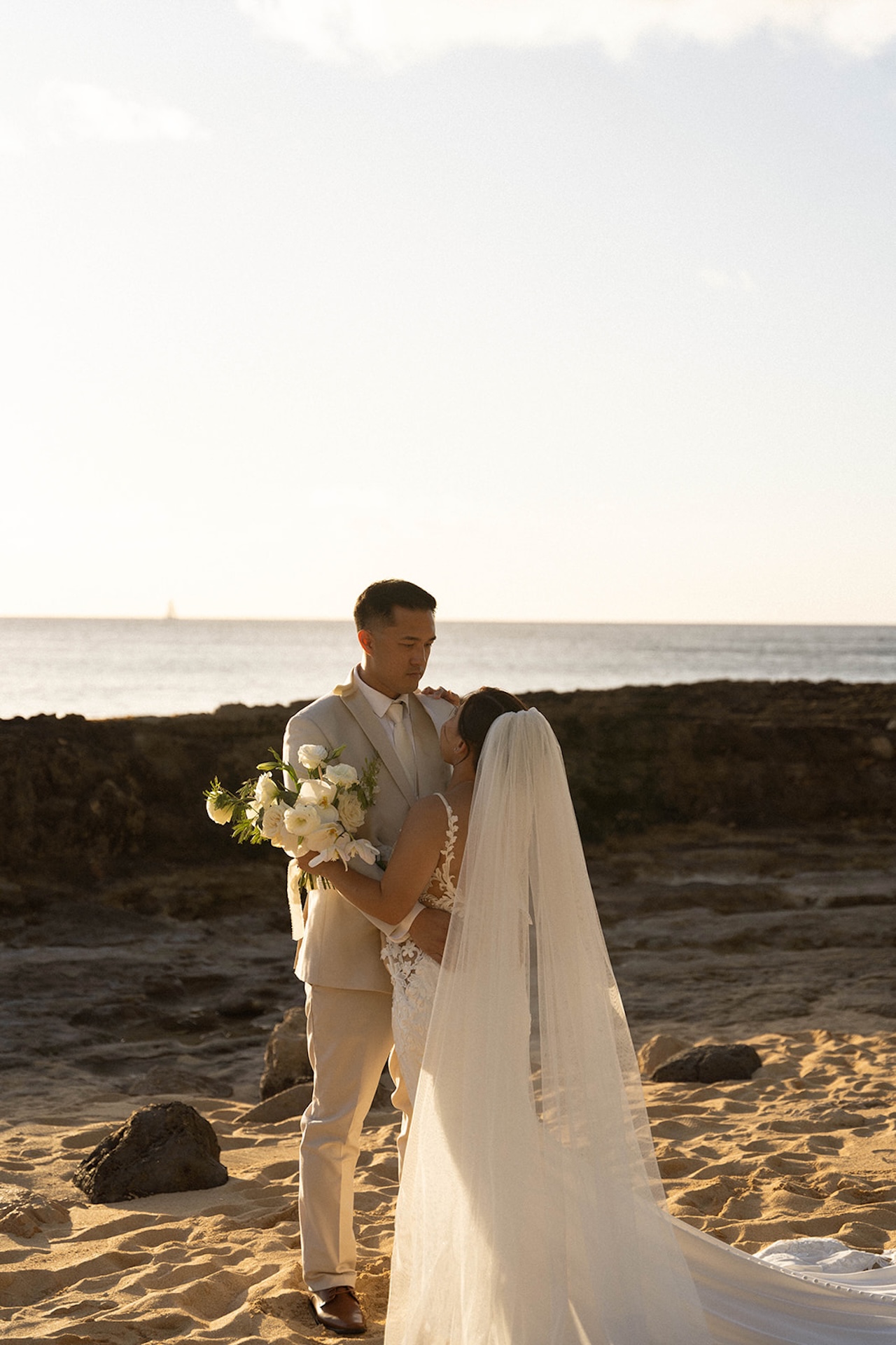 A bride and groom embrace on the beach at sunset, holding a bouquet of white flowers, a picture-perfect moment from their Destination wedding checklist.