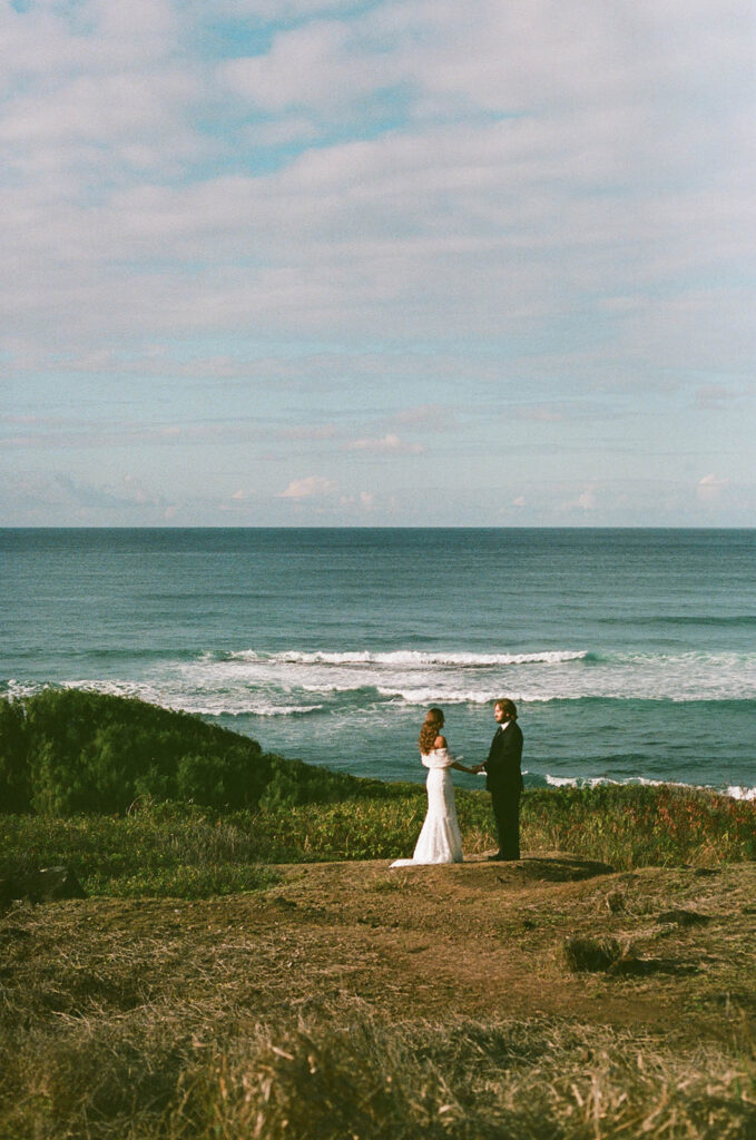 Bride and groom standing alone on a coastal overlook as they exchange vows with waves rolling in behind them, documented in a wide, atmospheric documentary wedding photography shot.