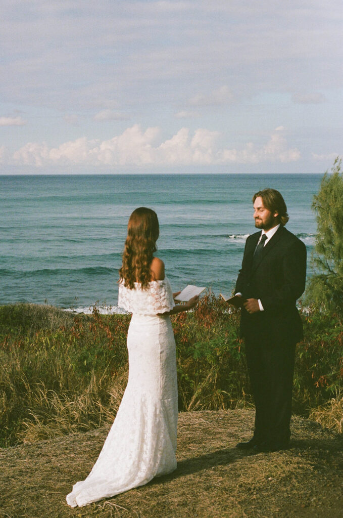 Bride and groom standing alone on a coastal overlook as they exchange vows with waves rolling in behind them.