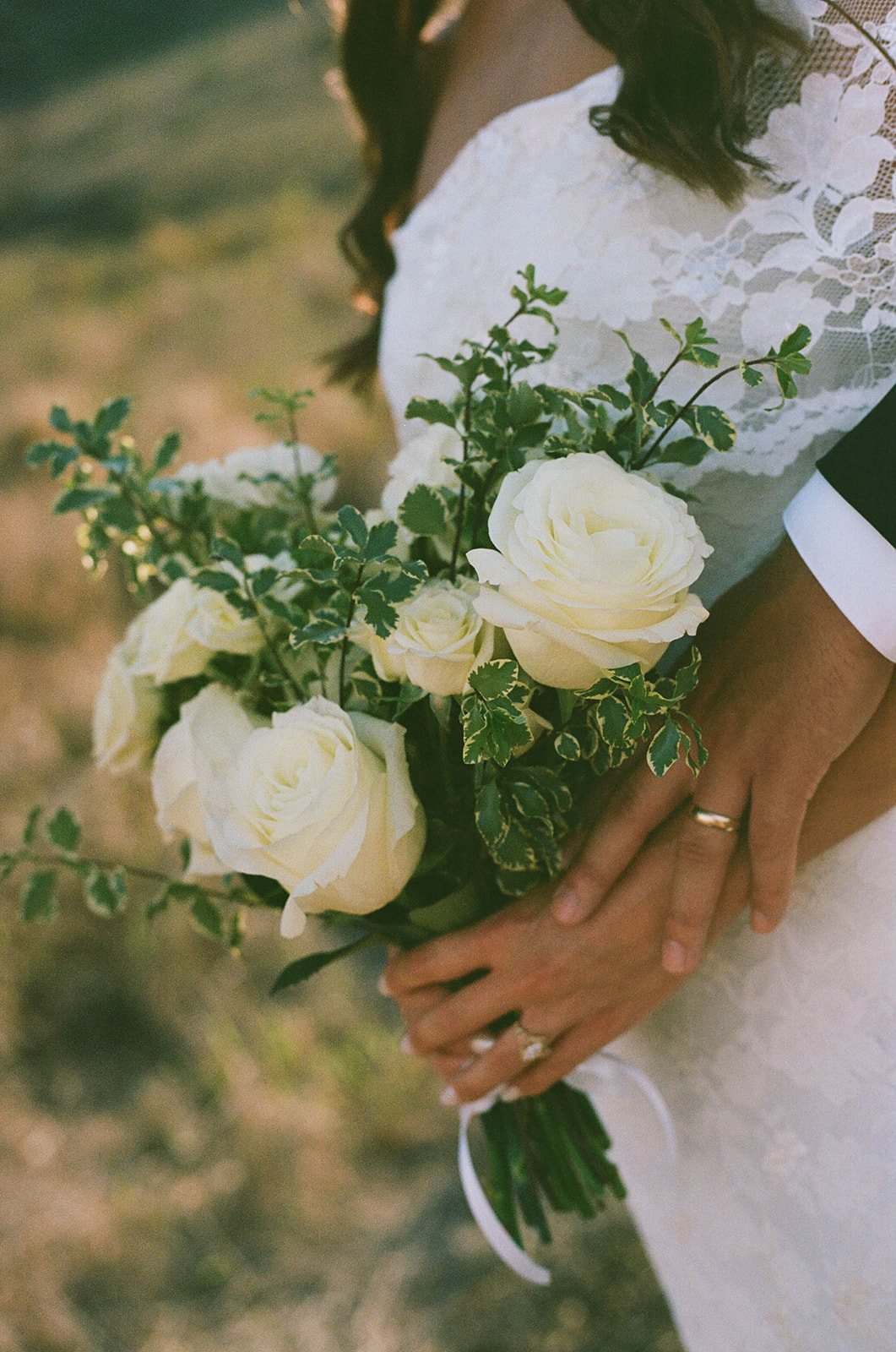 A close-up of the bride’s bouquet of white roses and greenery held between the couple’s intertwined hands, showing their wedding rings in a detailed documentary wedding photography shot.