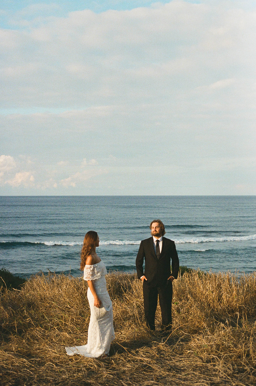 Couple standing in tall grass overlooking the water, a nostalgic film-style portrait from their Kauai Wedding.