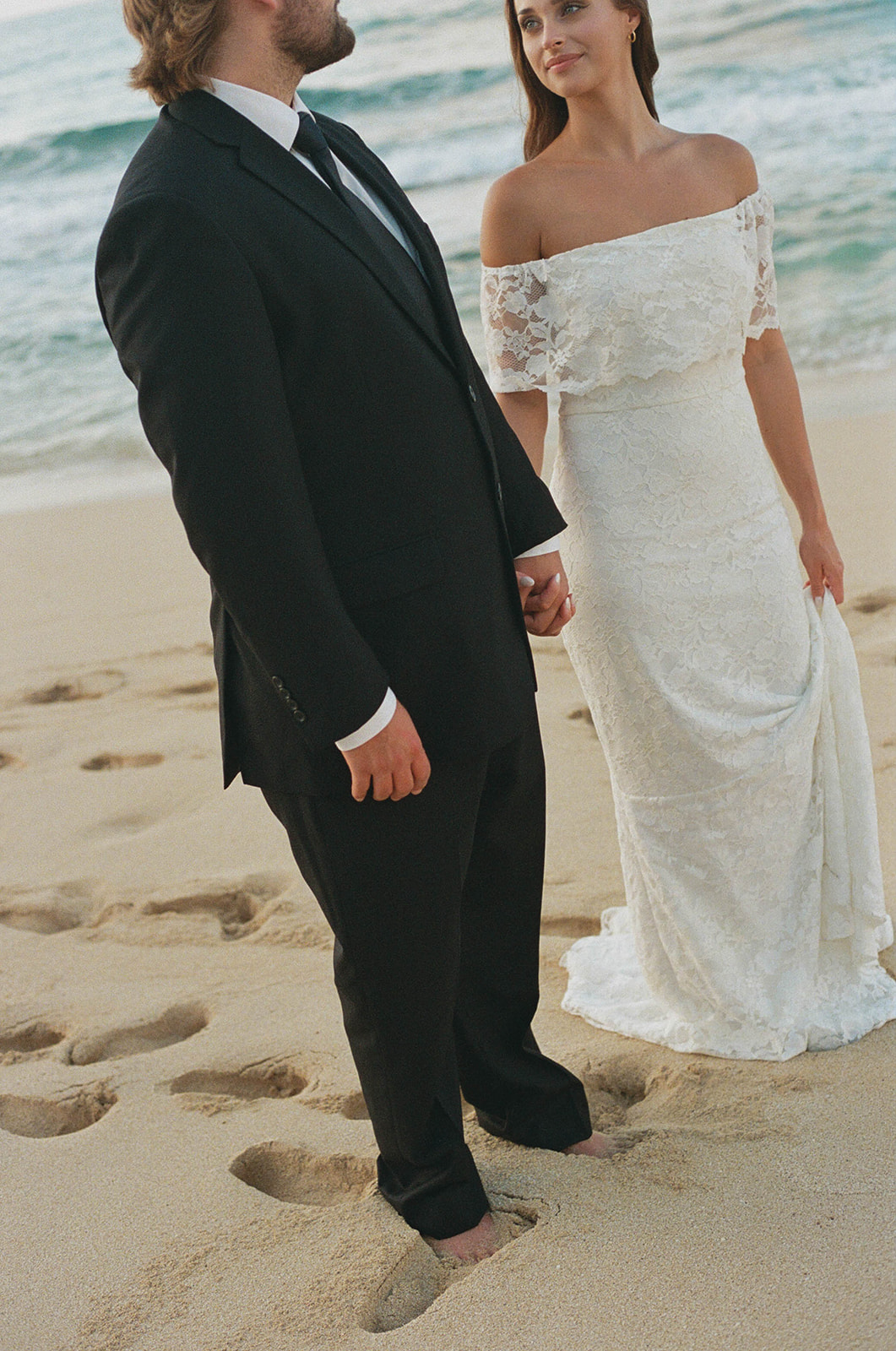 Groom in a black suit and bride in a lace off-the-shoulder gown holding hands on the sandy shore during their timeless Kauai Wedding Planning shoot.